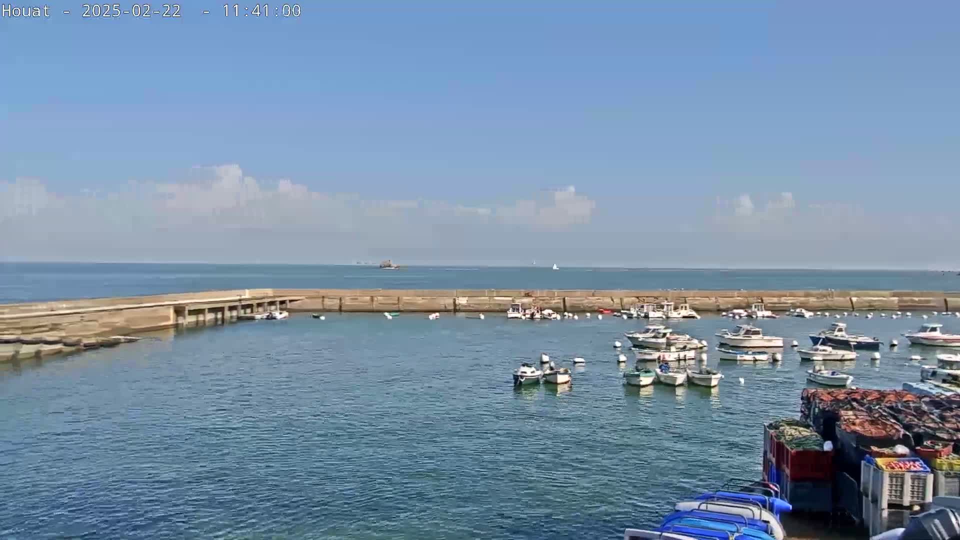 A calm, sunny day shows a harbor filled with numerous small boats moored within a stone seawall, with a clear blue ocean and a few clouds visible in the background.