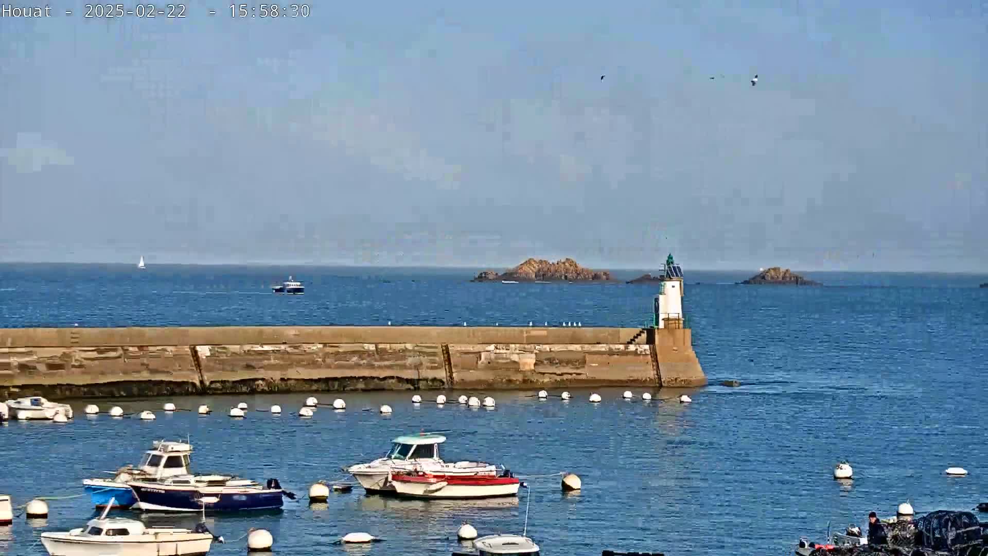 A harbor with several small boats moored alongside a stone wall, a small lighthouse, and a rocky island visible in the distance on a clear, sunny day.