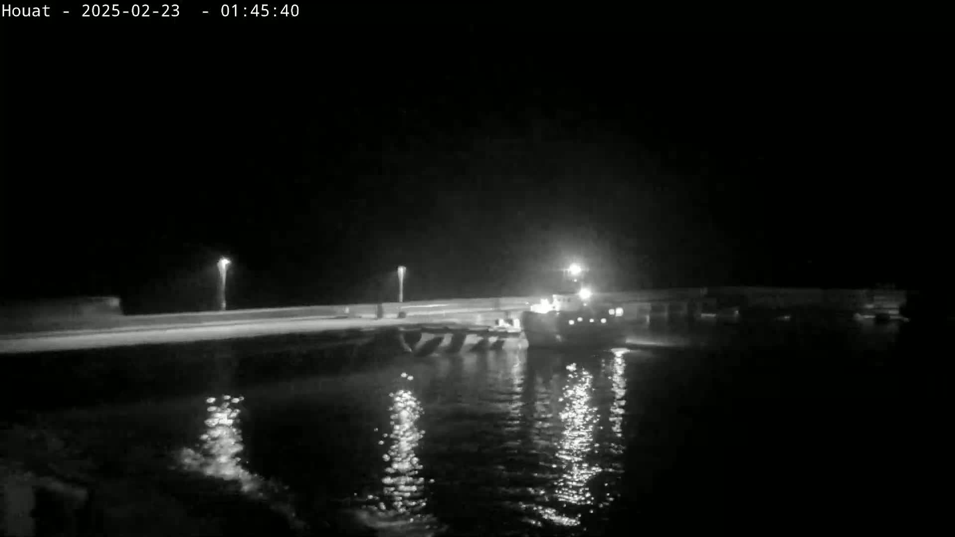 A nighttime, low-light view of a boat docked at a pier, with calm water reflecting lights.