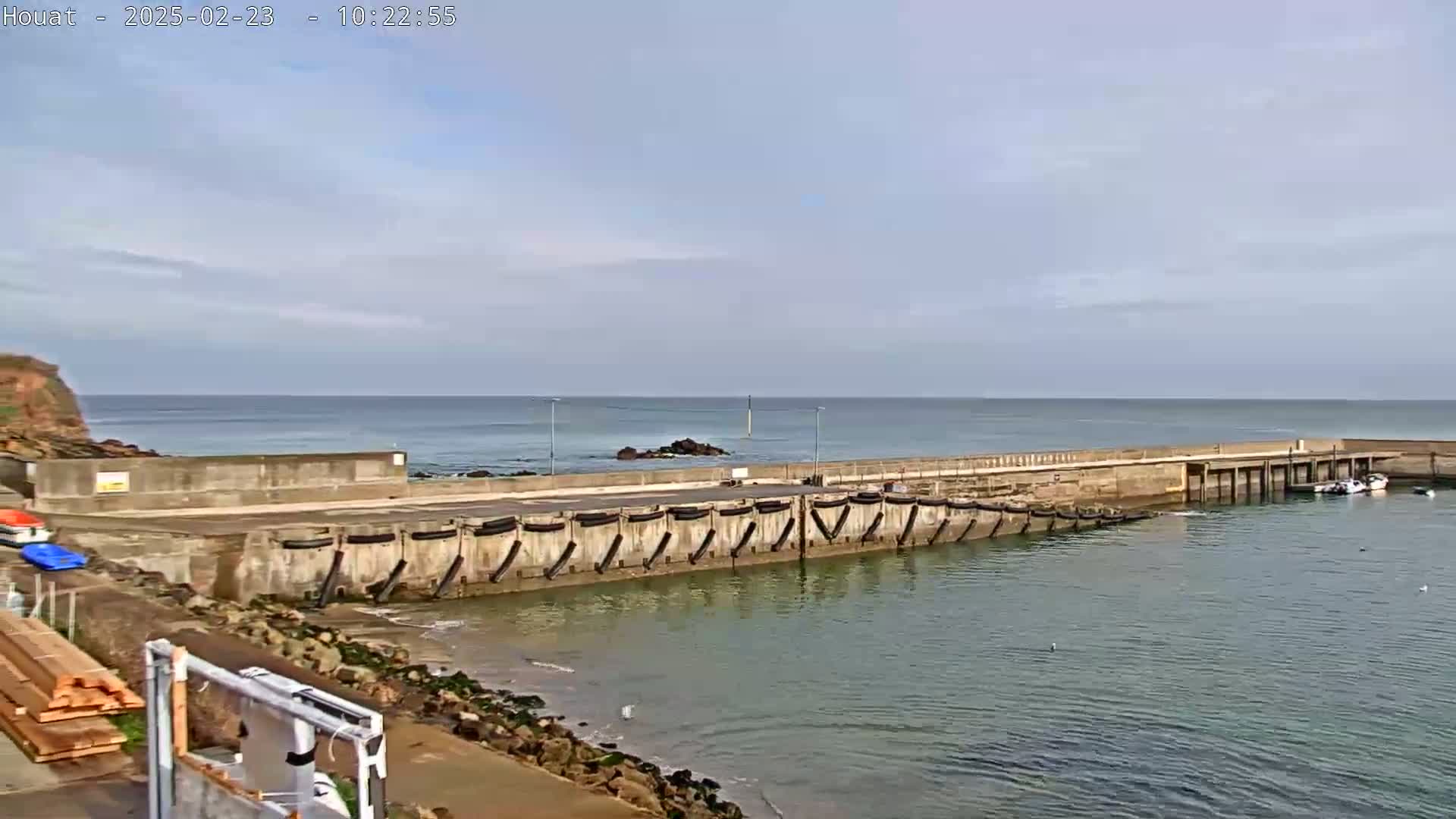 A concrete harbor wall extends into calm, light-blue ocean water under a mostly clear sky.