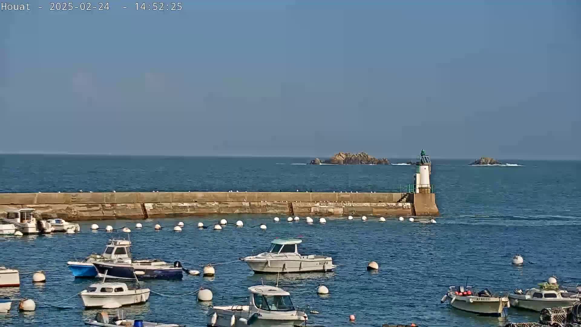A harbor filled with small boats sits beside a low stone wall, with a small lighthouse at the end and calm blue ocean under a clear sky.