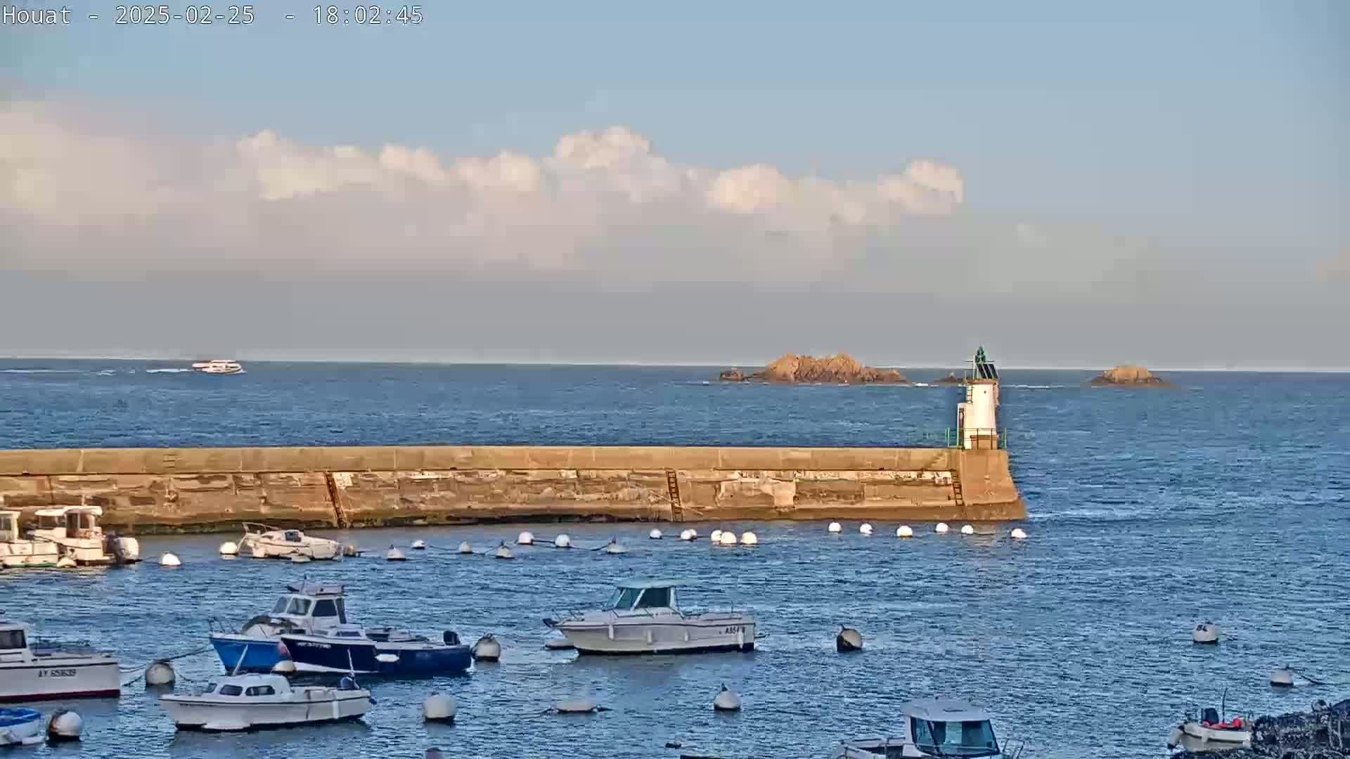 A calm sea with partly cloudy skies shows a small harbor with several moored boats and a lighthouse at the end of a seawall, with distant rocky islands.