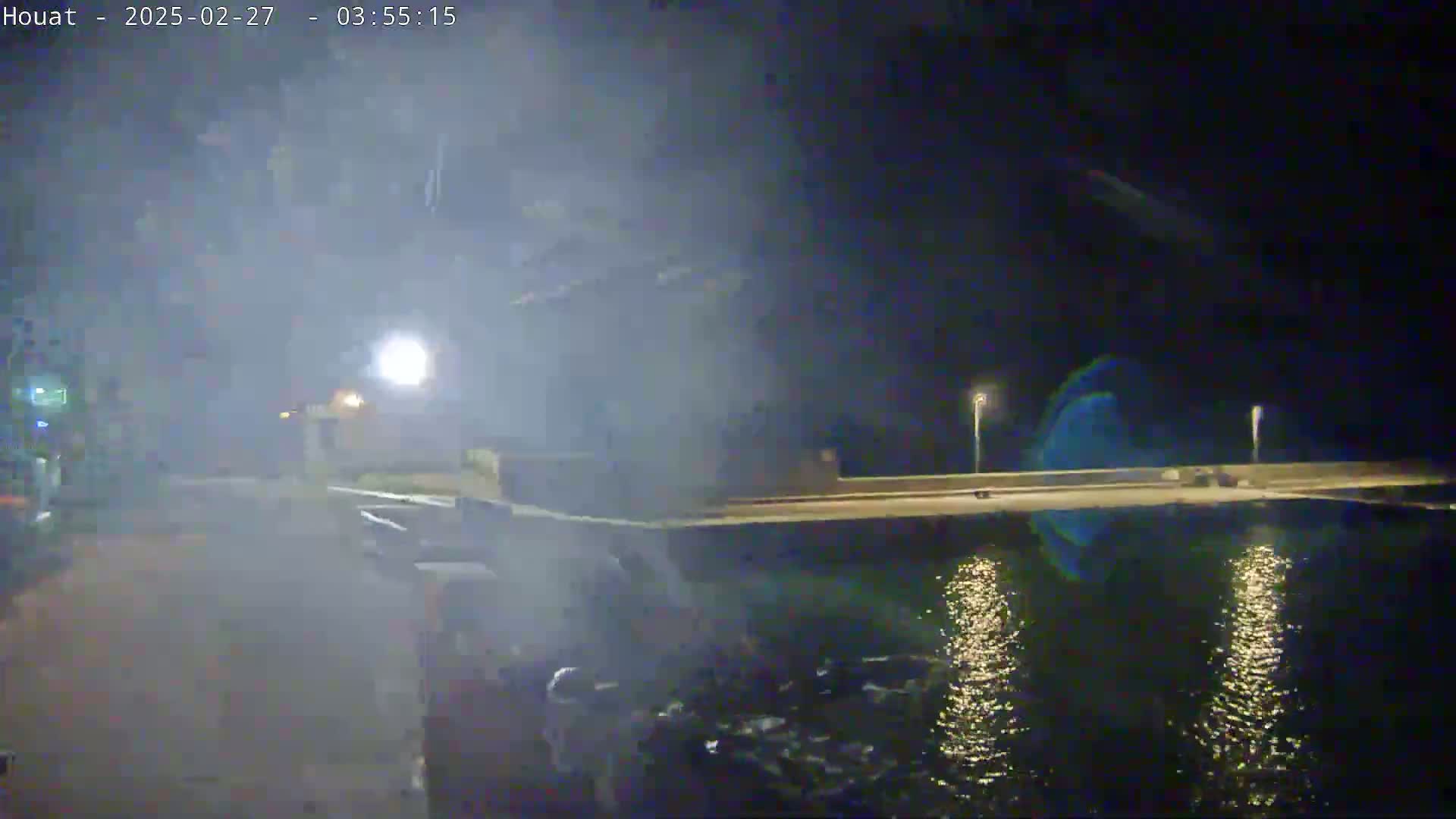 A nighttime view of a hazy waterfront scene shows a roadway leading to a low wall beside calm water reflecting lights.