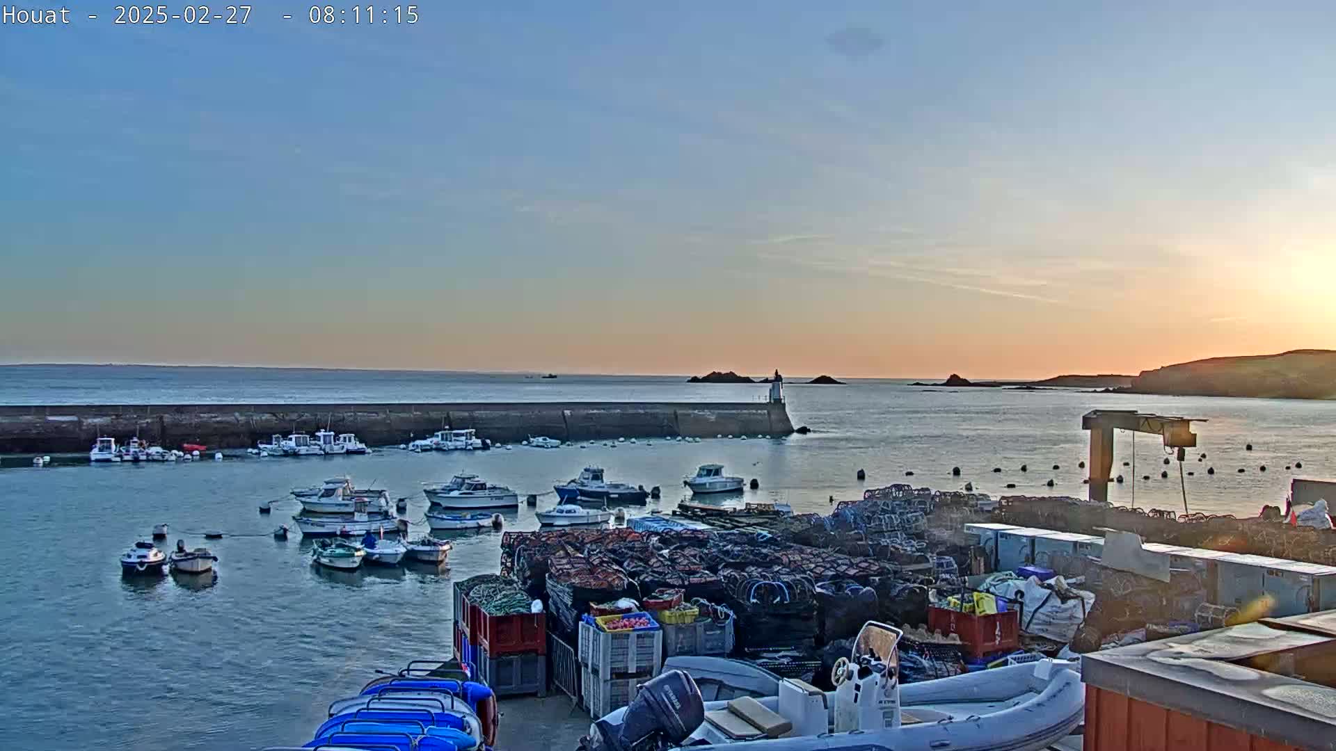 Many small boats are moored in a calm harbor at sunset, with a stone seawall and stacks of fishing equipment visible on the shore.