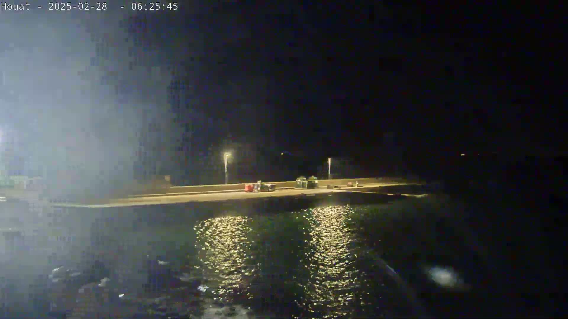 A nighttime view of a dark, calm body of water reflecting the lights from a nearby dock where several vehicles are parked.