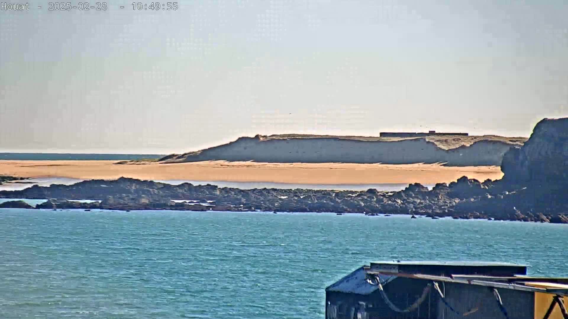 A sandy beach backed by a rocky cliff is visible across a calm body of blue water, with a portion of a dark structure in the foreground.