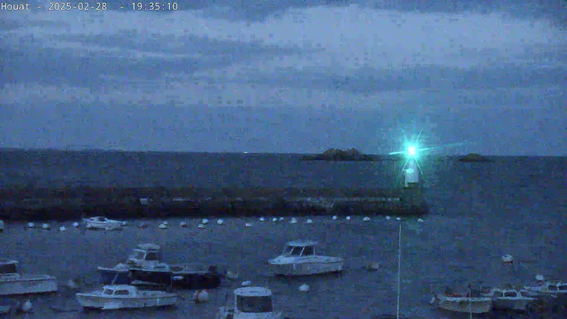 Under a twilight sky, numerous small boats are moored in a harbor next to a lit lighthouse, with a few small islands visible in the distance.