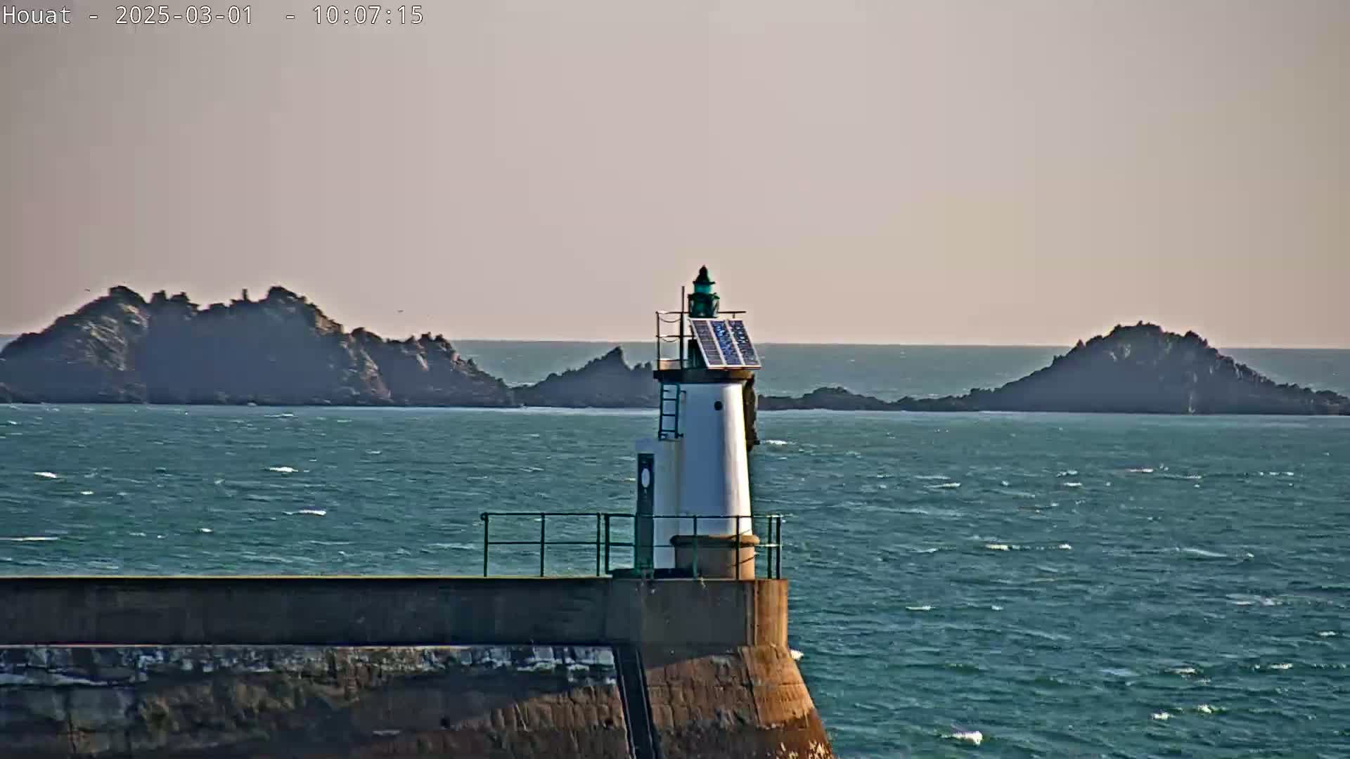 A small, solar-powered lighthouse stands atop a seawall overlooking a choppy sea and distant rocky islands under a mostly clear sky.