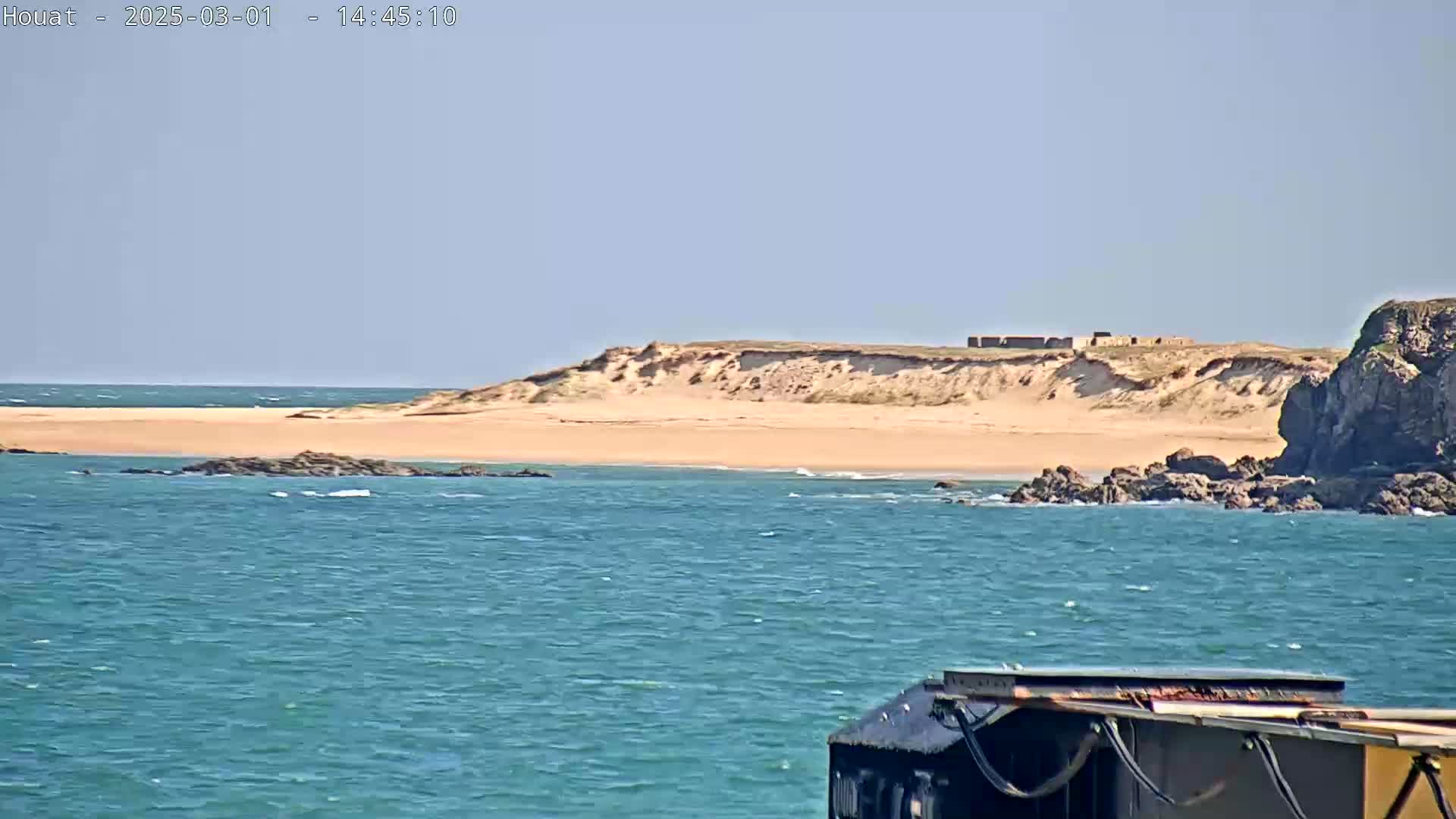 A sandy beach backed by dunes and a rocky outcrop is visible across a body of teal water, under a clear sky.