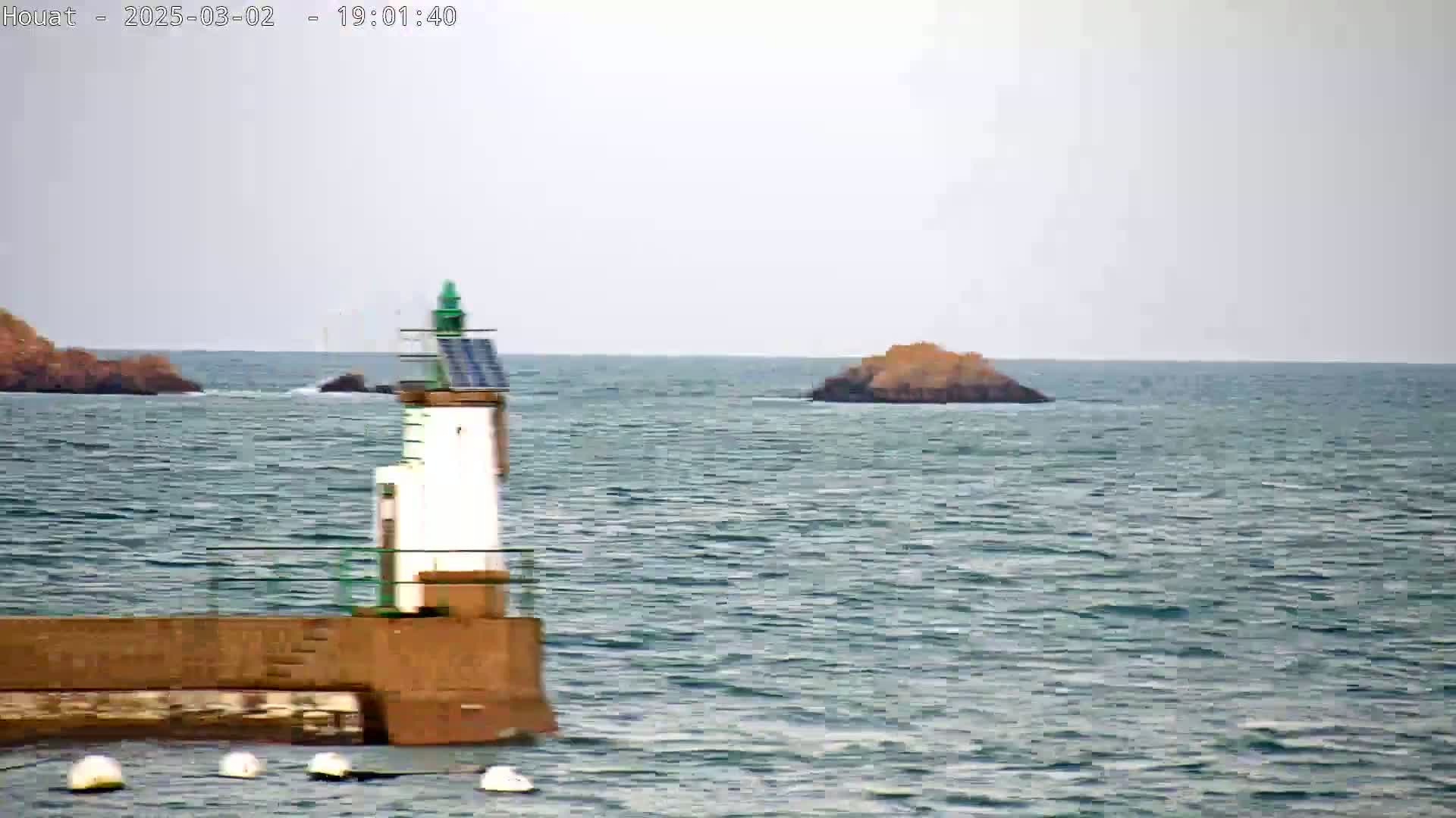 A small lighthouse with a solar panel sits on a brown wall, overlooking a choppy sea with two small islands in the distance under an overcast sky.