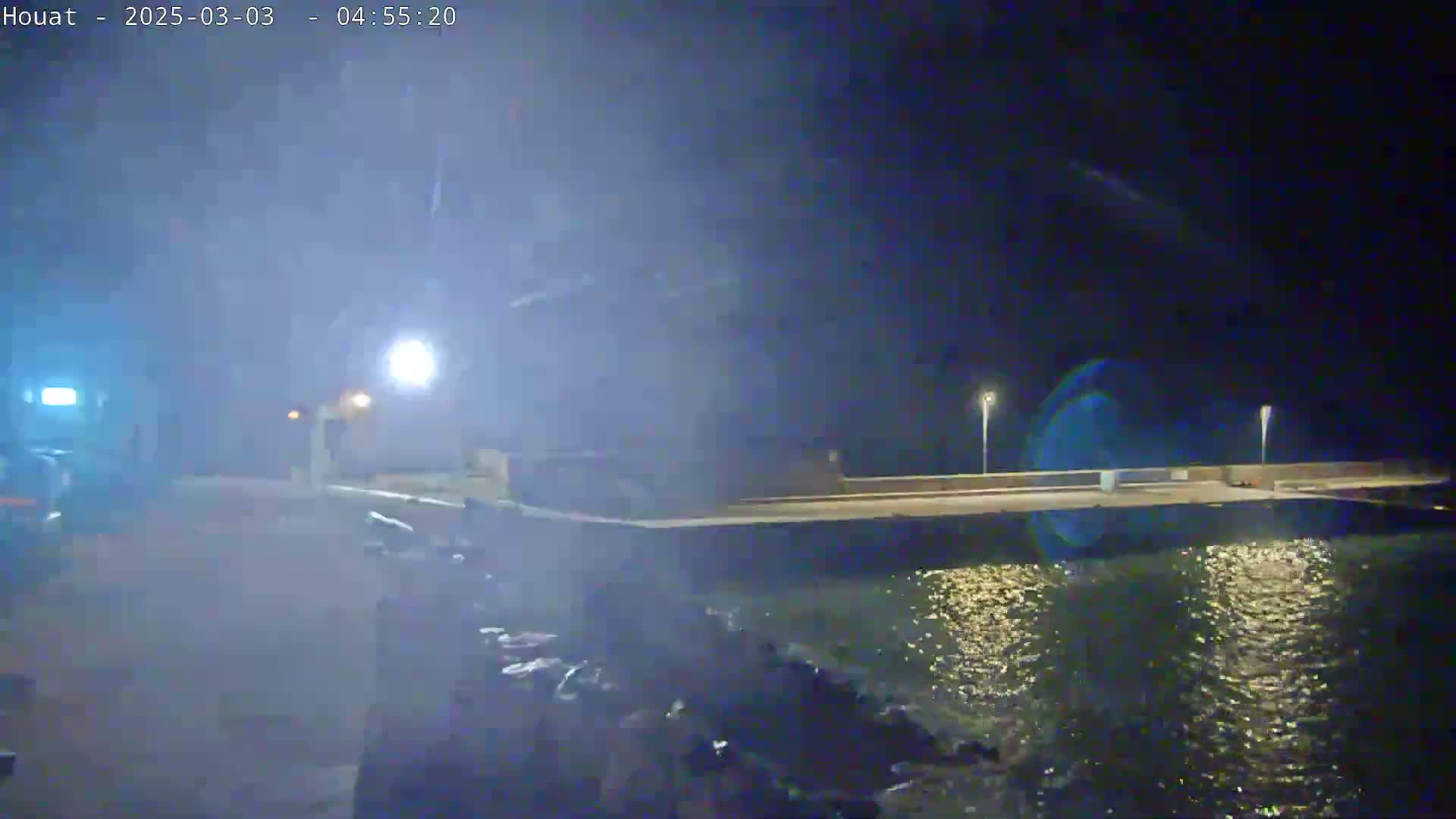 A nighttime view of a waterfront area with a low wall separating land and water under misty or foggy conditions.