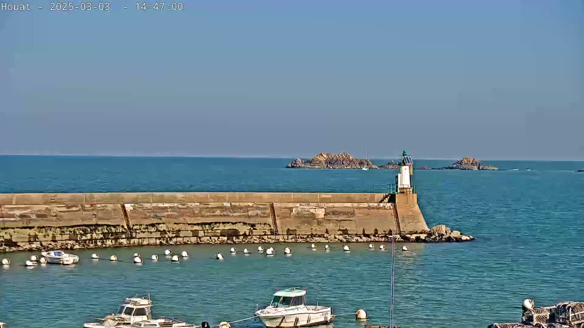 A harbor with several small boats is protected by a long stone wall and a small lighthouse, all under a clear, sunny sky.