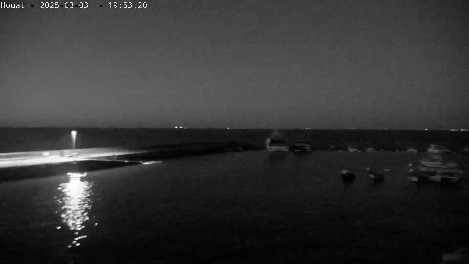 A nighttime, low-light view of a calm body of water with several boats moored near a shoreline, which is illuminated by a streetlight.