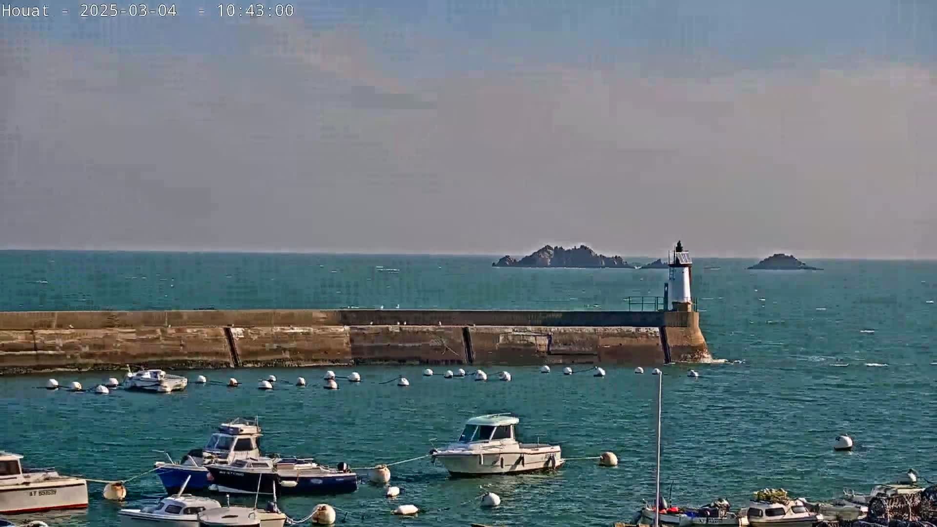 A harbor with several small boats is protected by a seawall and features a lighthouse, with small islands visible in the distance on a calm, sunny day.