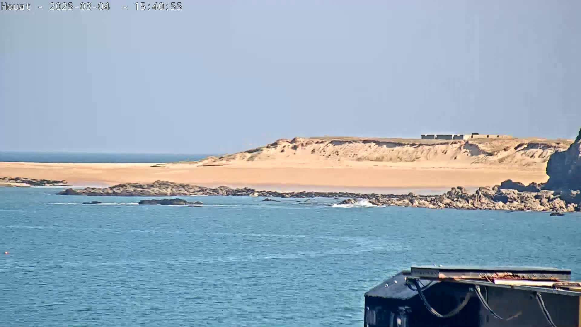 A sandy beach and rocky shoreline are visible across a calm blue body of water, under a clear sky.