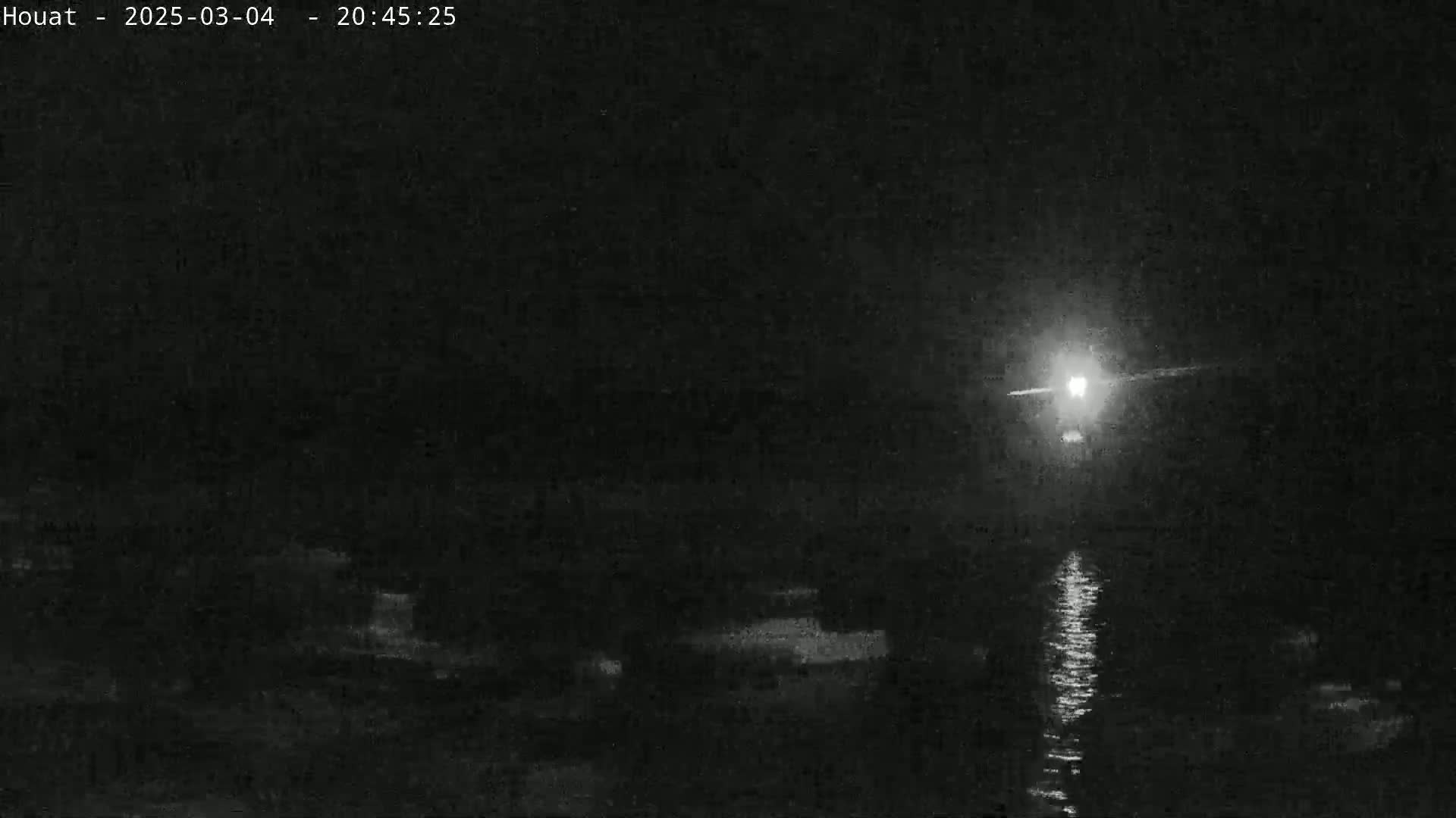 A bright light shines over dark water at night, reflecting on the surface, with a dark shoreline visible in the foreground.