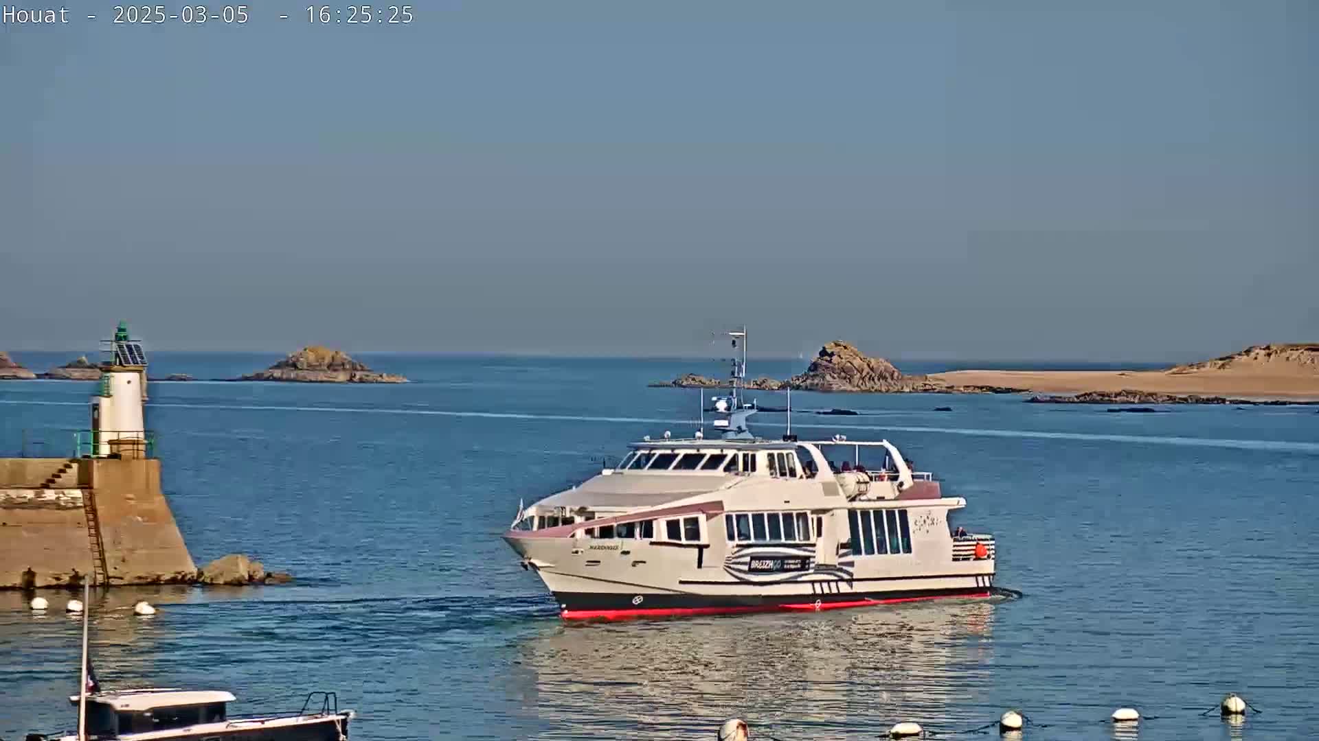 A ferry approaches a harbor on a calm, sunny day, passing a lighthouse on a stone jetty with small islands in the background.