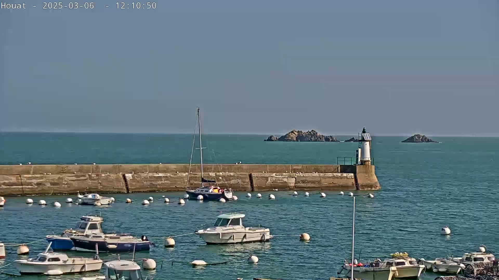 A harbor filled with moored boats is situated next to a stone seawall with a lighthouse at its end, under a clear blue sky and calm sea with small islands visible in the distance.