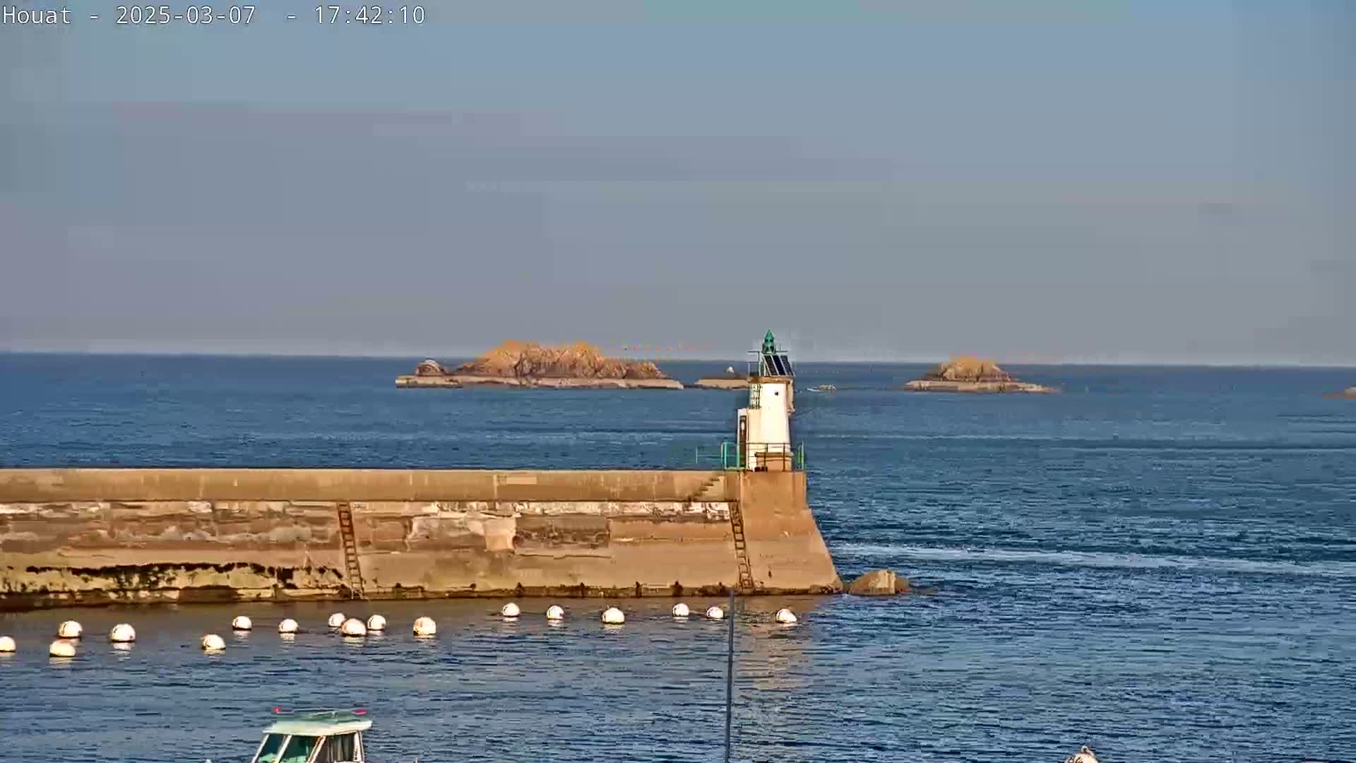 A small lighthouse sits atop a seawall in calm water, with several small islands visible in the distance under a clear sky.