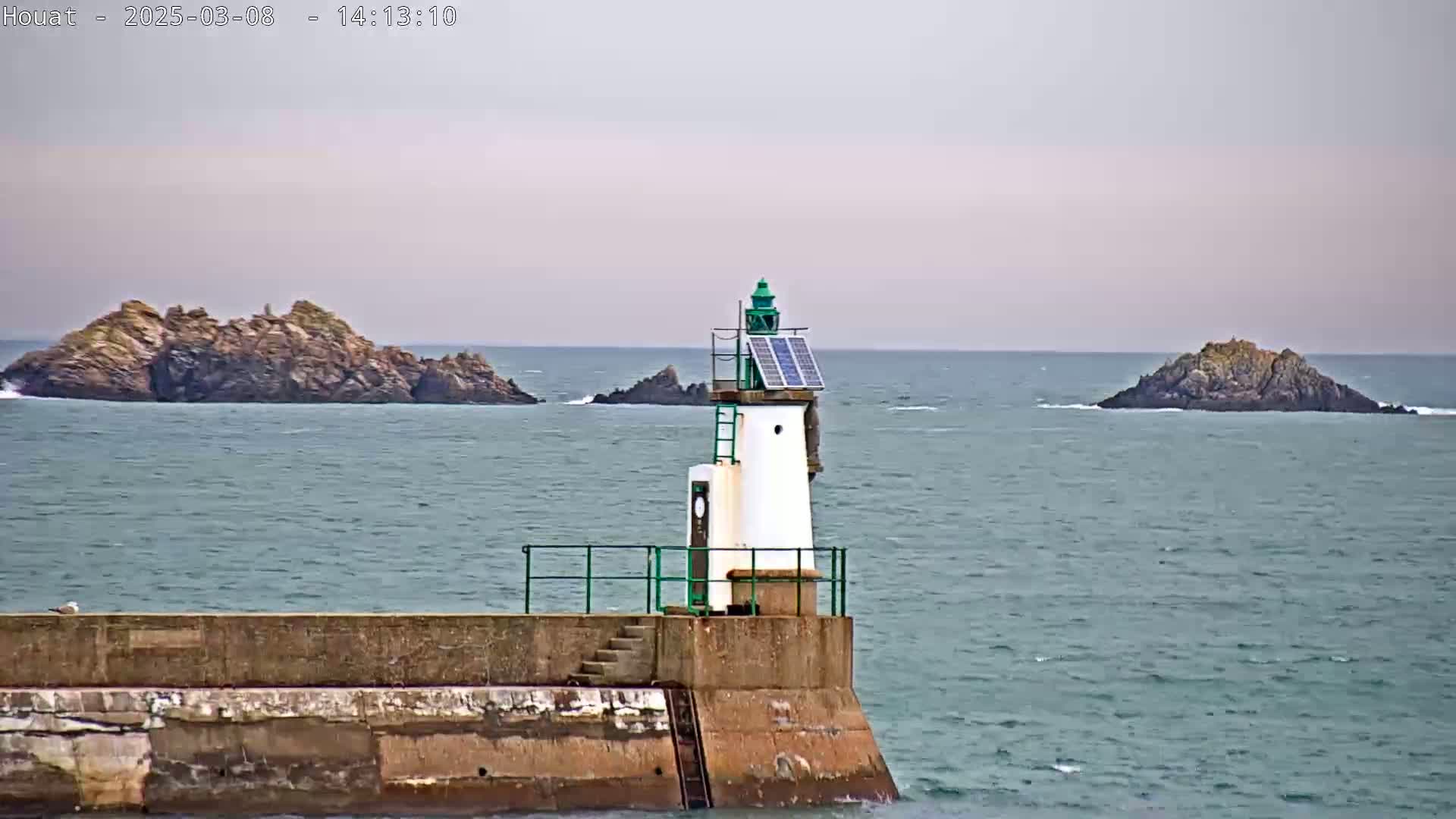 A small, solar-powered lighthouse stands on a seawall, overlooking a calm sea with rocky islands in the distance under an overcast sky.