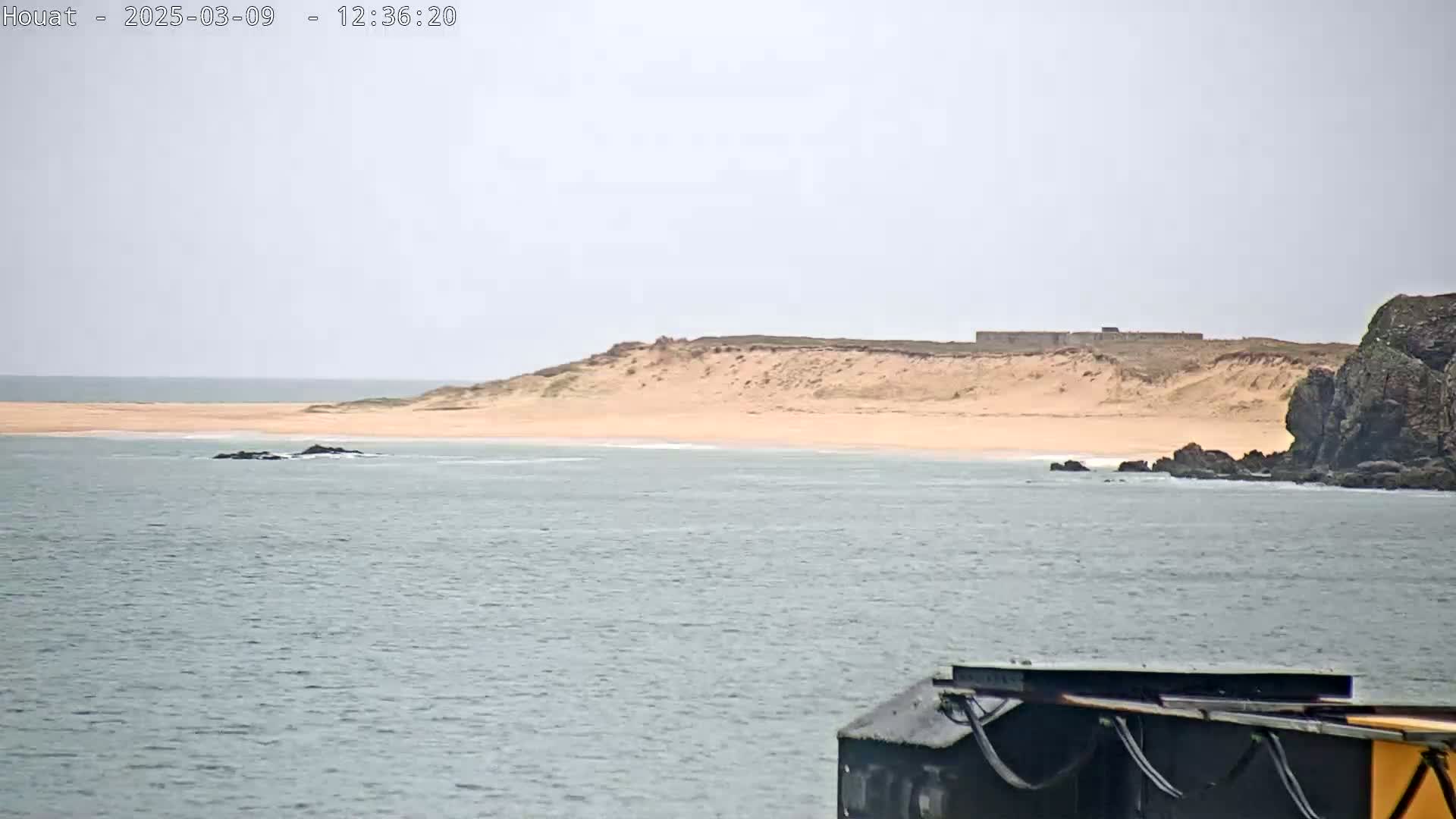 A sandy beach backed by dunes and some structures is visible across a calm body of water from a dark-colored structure in the foreground.