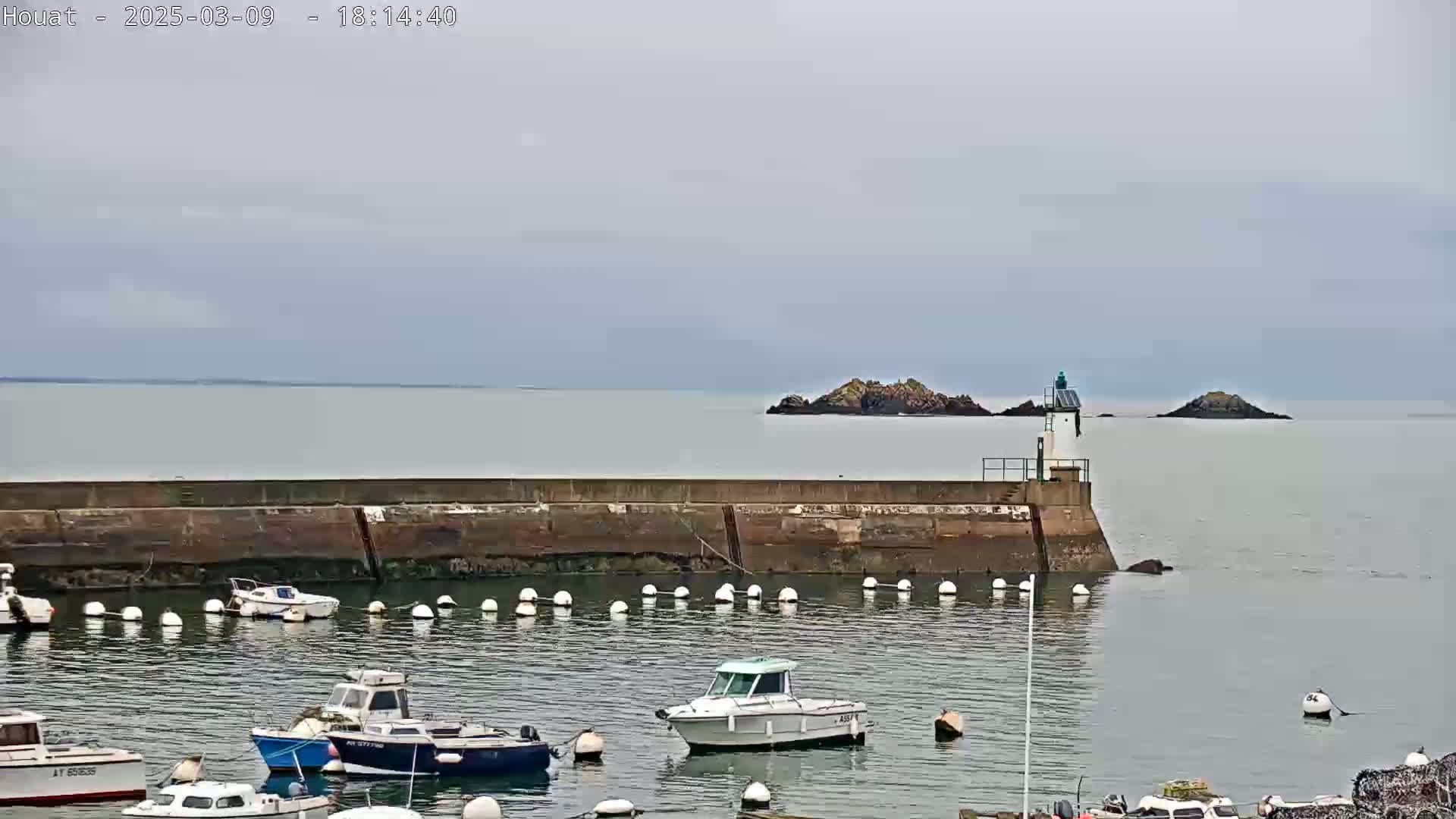 A calm, overcast day reveals a harbor filled with small boats, protected by a concrete wall and a lighthouse, with small islands visible in the distance.