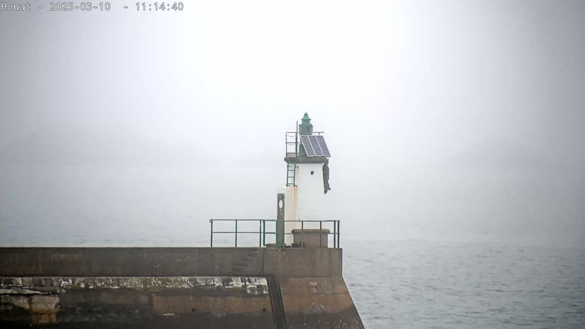 A small lighthouse with a solar panel sits atop a seawall on a foggy day.