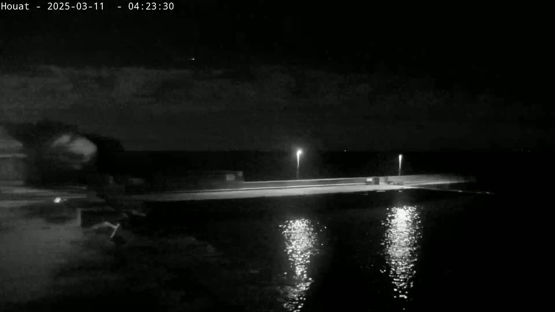 A nighttime, low-light view shows a long, low structure extending over dark water, illuminated by two lights; the sky is mostly cloudy.
