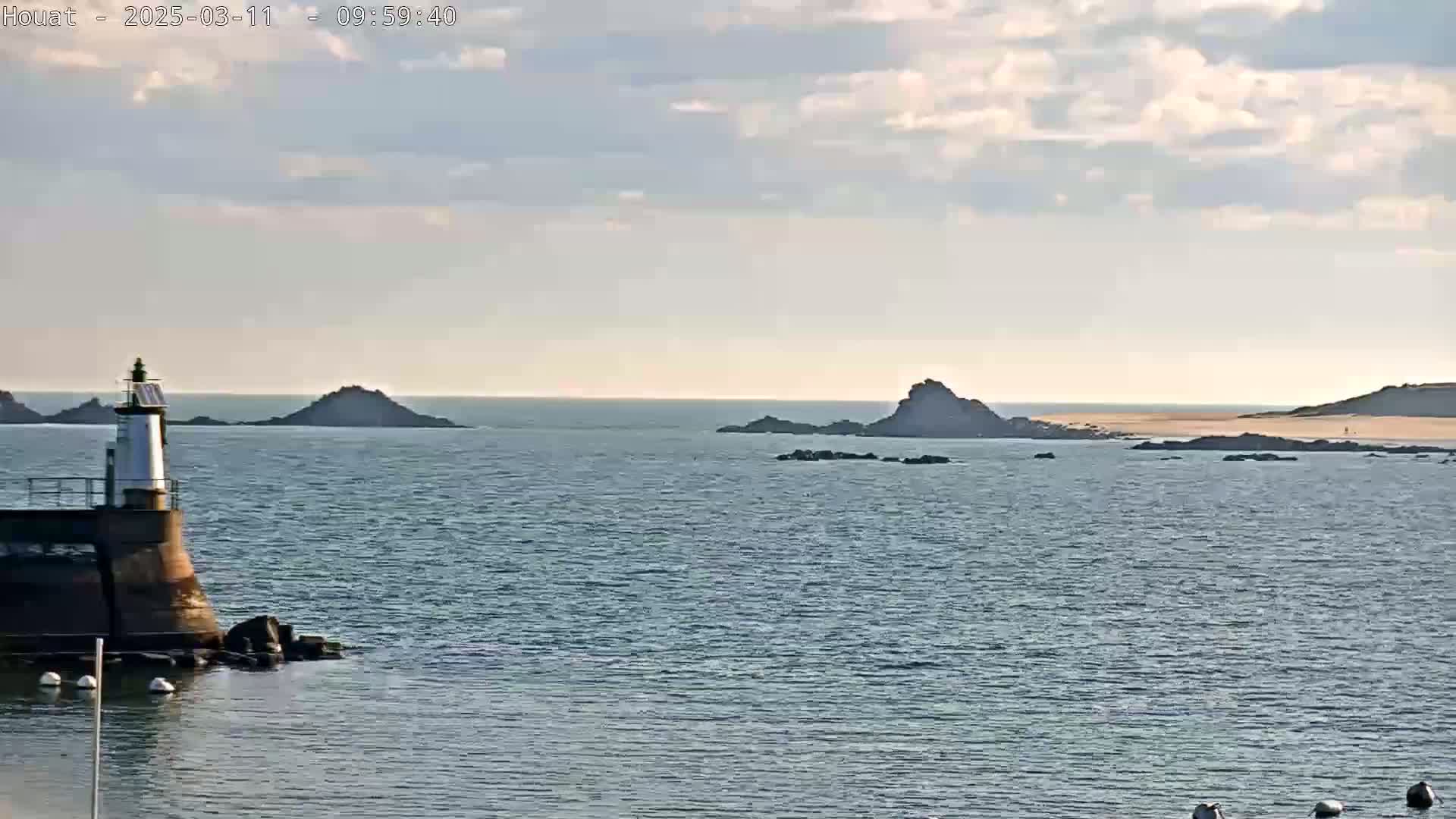 A lighthouse stands on a rocky outcropping near a calm ocean under a partly cloudy sky.