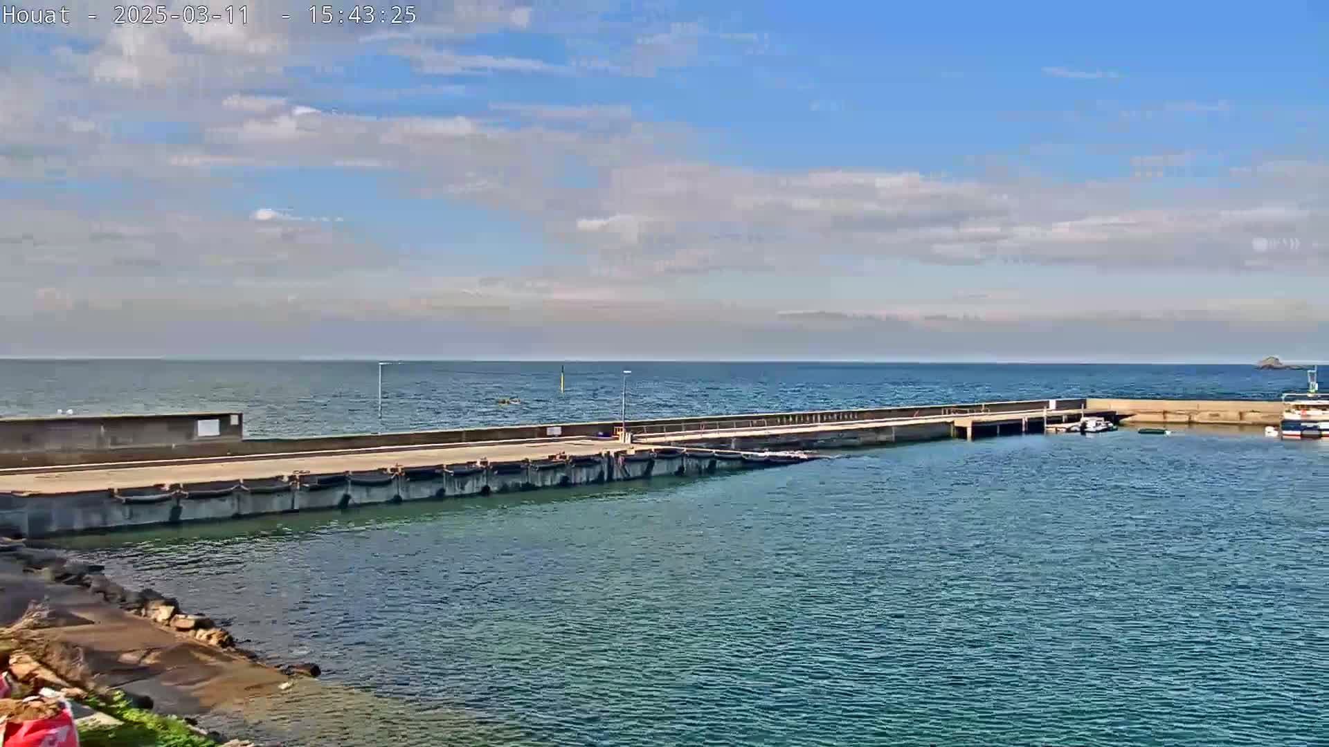 A concrete pier extends into a calm, blue ocean under a mostly sunny sky with scattered clouds.