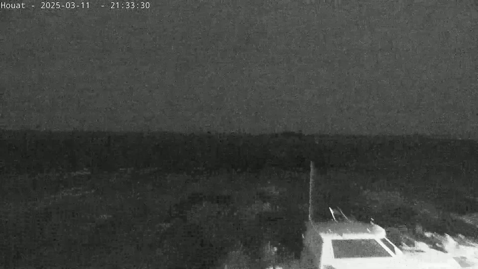 A nighttime, low-light view shows dark land and a portion of a vehicle's roof in the bottom right corner under a mostly clear sky.