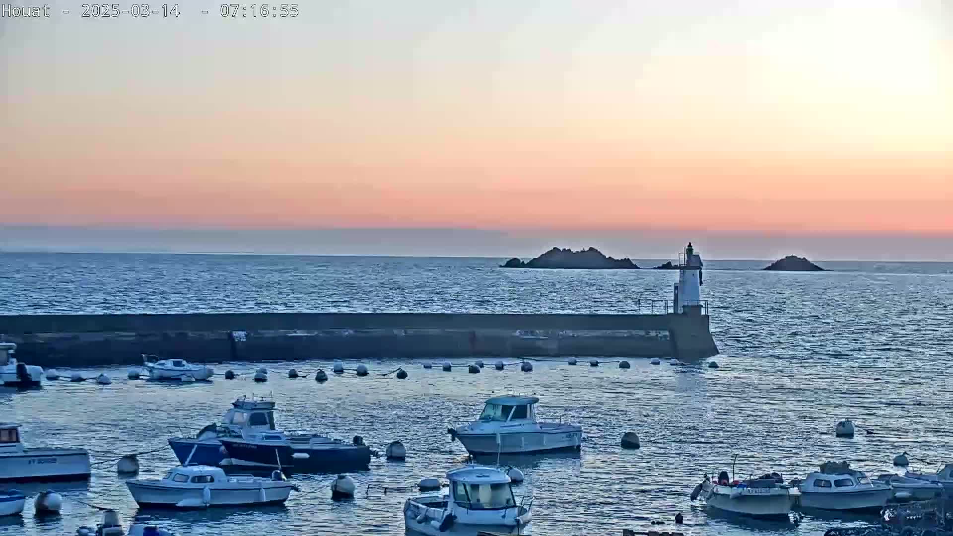A calm harbor at sunset, filled with small boats, is protected by a seawall, with a small lighthouse and distant islands visible under a pale pink sky.