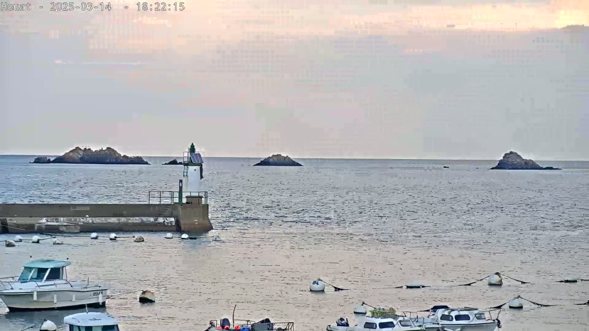 A small harbor with several boats is situated near a lighthouse, overlooking a calm sea with several small rocky islands in the distance under a cloudy, overcast sky.