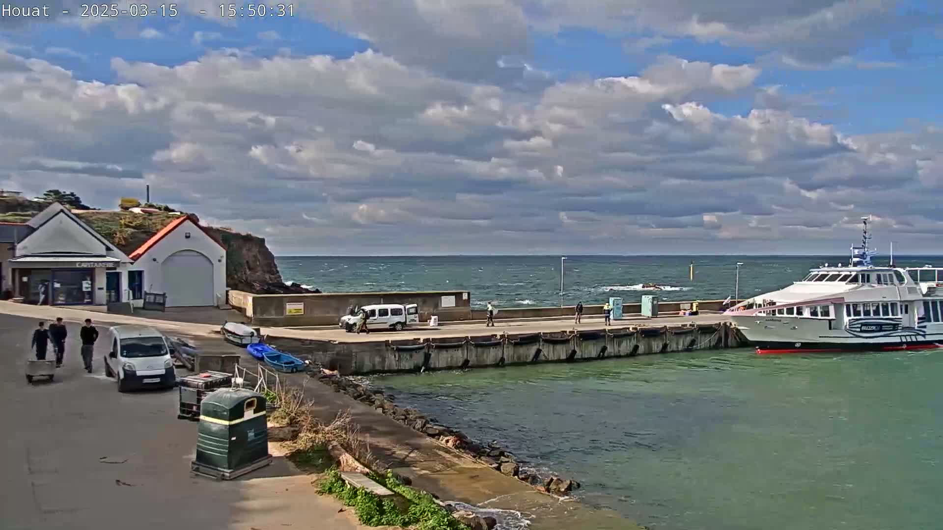 A ferry is docked at a harbor, with several people walking near a small building and vehicles on a partly cloudy day with a windy sea.