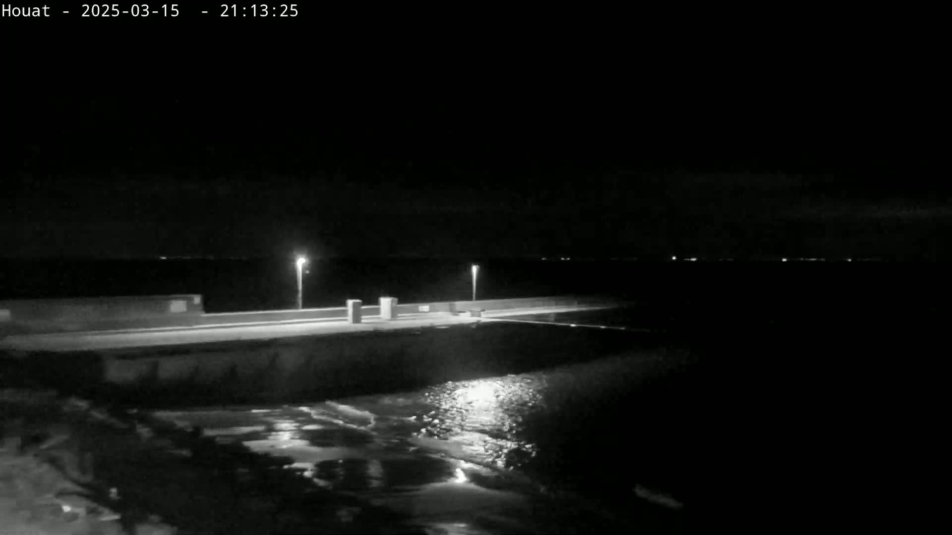 A nighttime, low-light image shows a dark, calm body of water lapping at a low seawall illuminated by several lights.