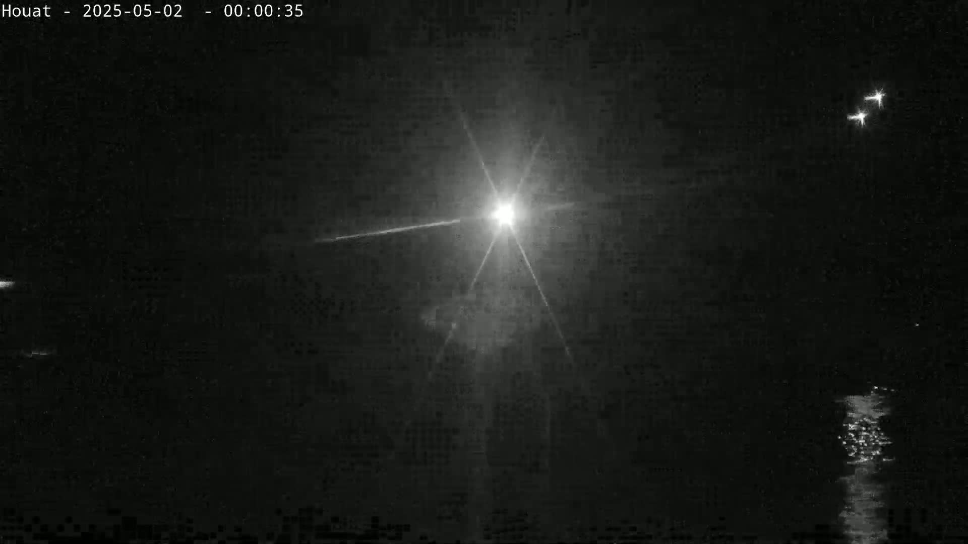 A bright light, possibly a spotlight or searchlight, shines across a dark nighttime scene with water visible in the lower right corner and faint lights in the distance.