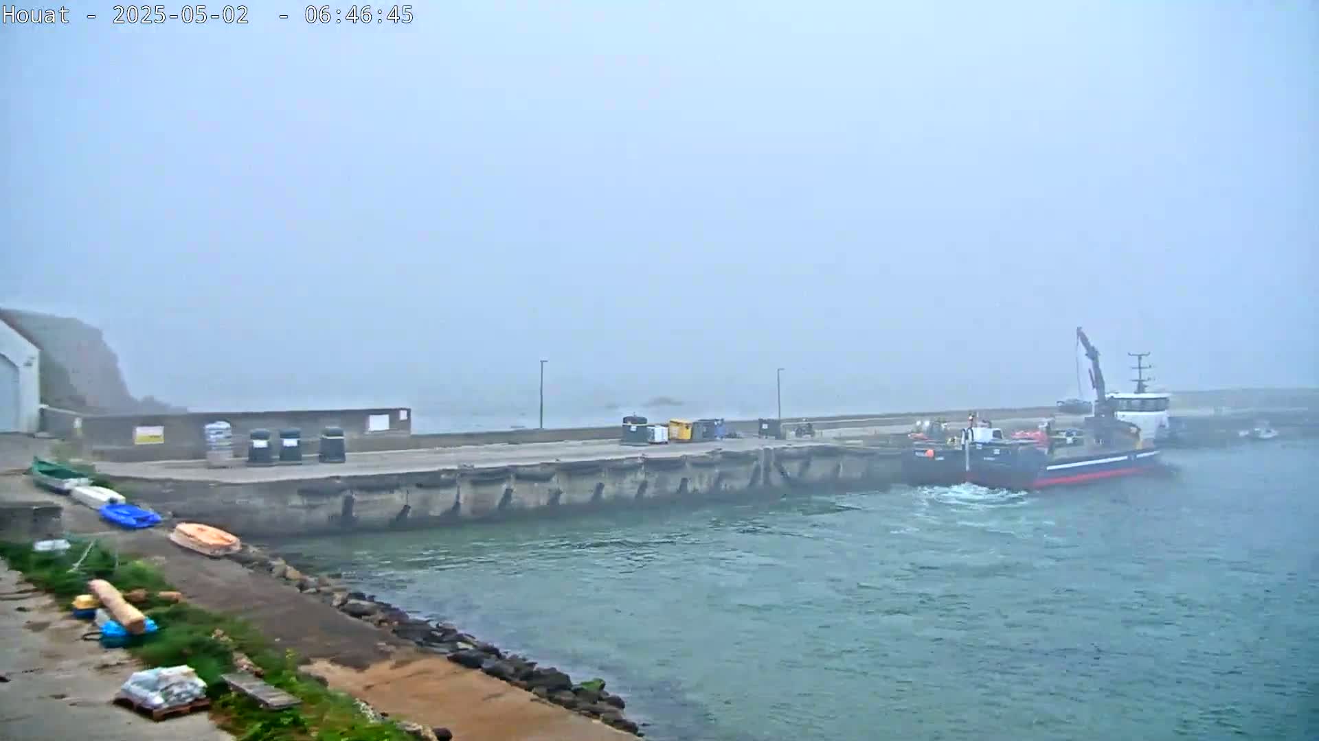 A small harbor in foggy conditions shows a crane barge docked next to a concrete wall, with several smaller boats and some debris along the shore.