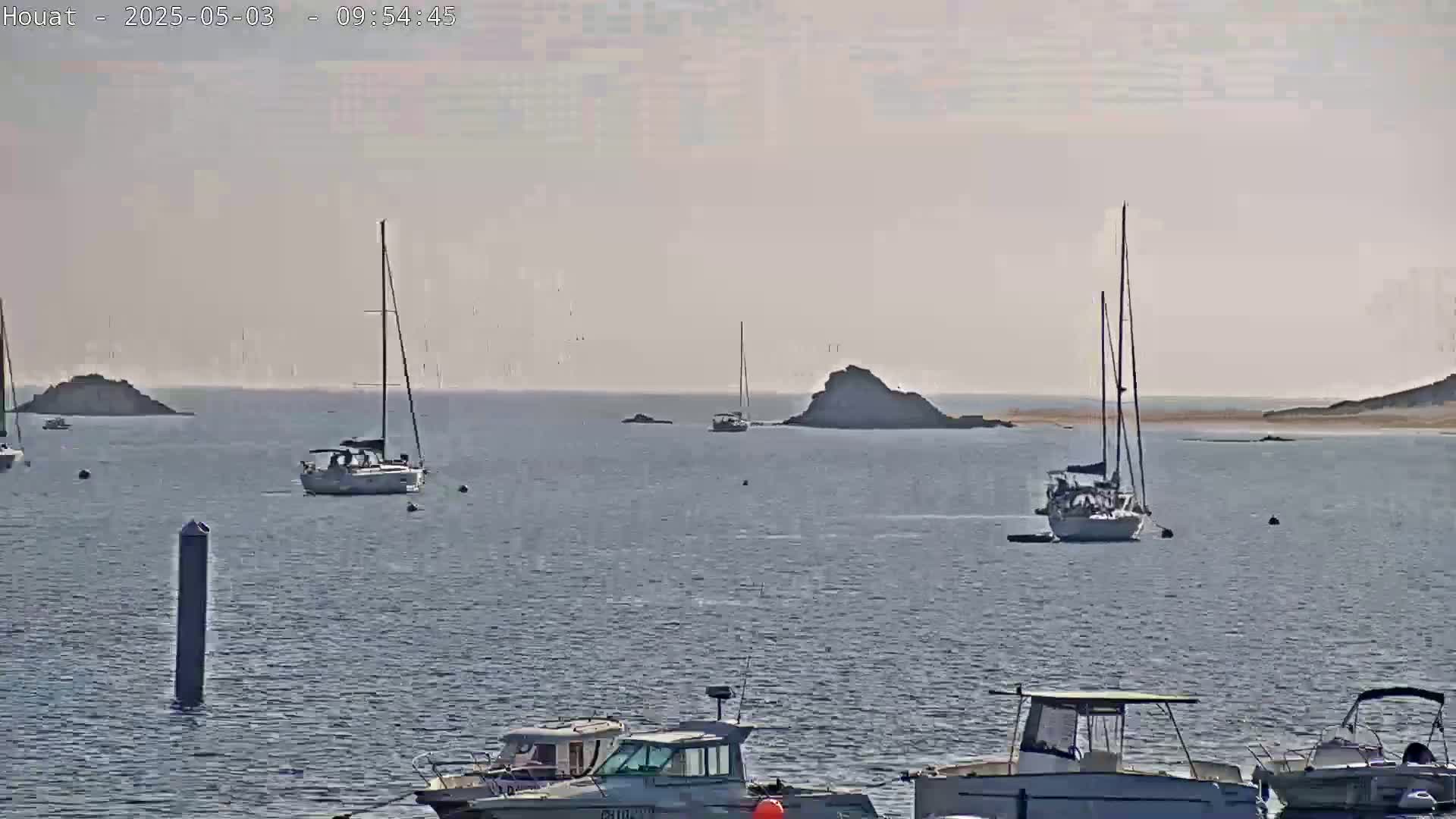 Several sailboats and motorboats are at anchor in calm, hazy waters near some small rocky islands.