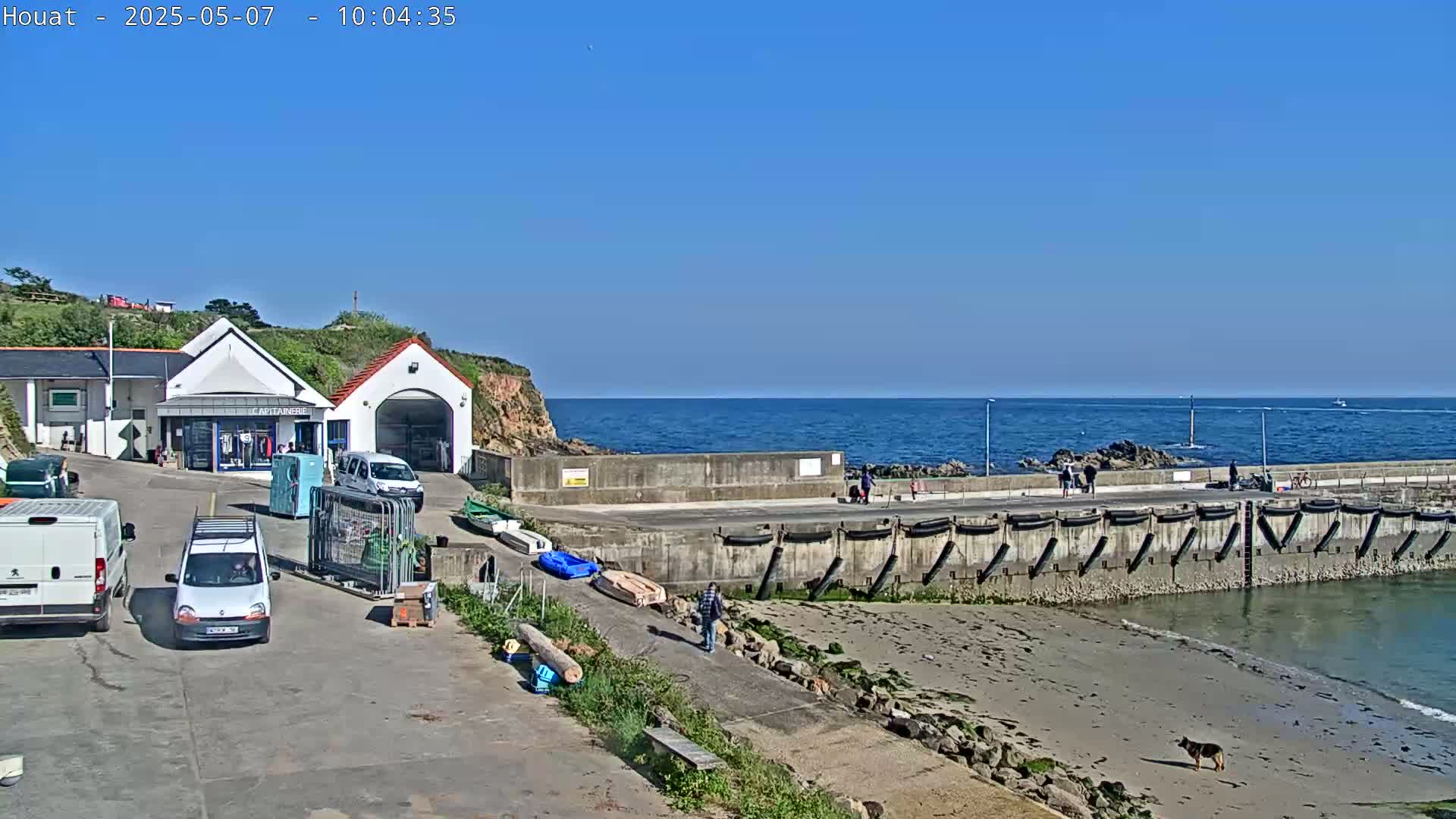 A small harbor with a building, several vehicles, and a few people is visible on a sunny day, with a calm ocean in the background.