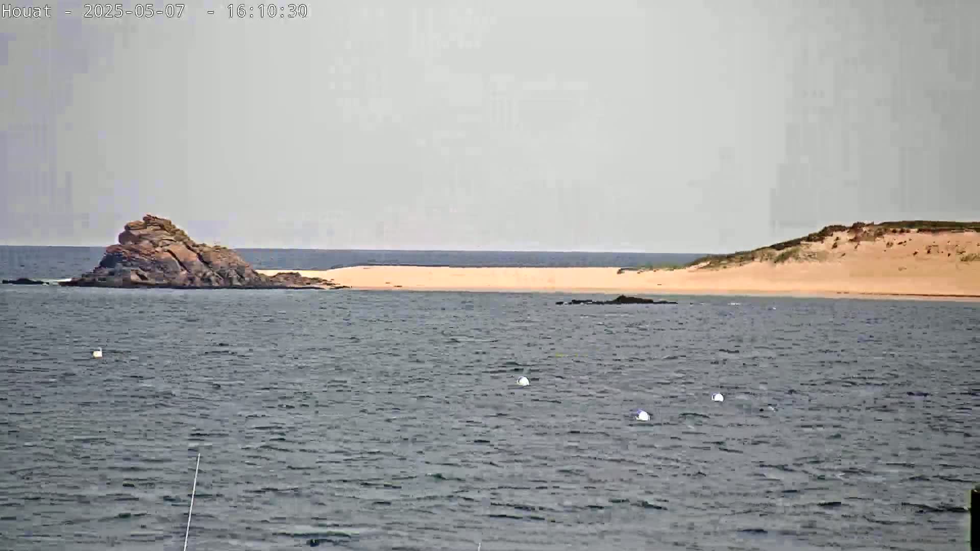 A rocky islet and a sandy expanse of land are visible across a body of water under an overcast sky.