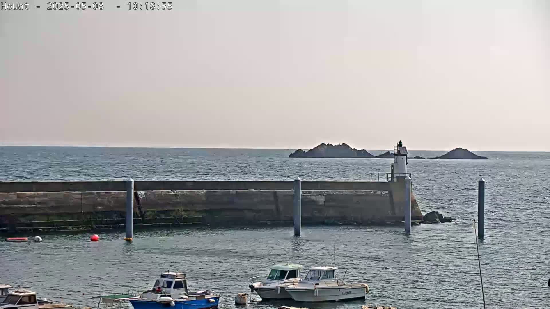 A harbor with several small boats is partially protected by a seawall, beyond which is a calm sea, two small islands, and a lighthouse.