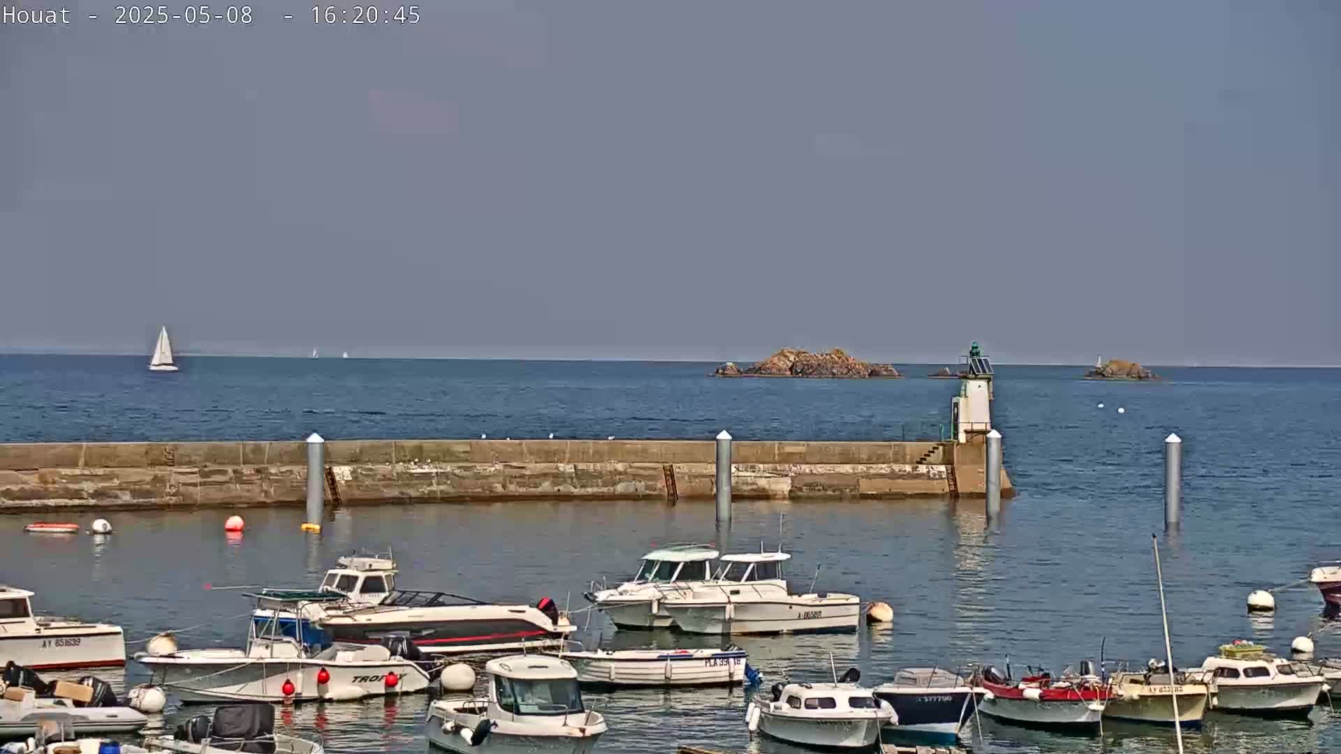 A calm sea with a few sailboats in the distance is seen behind a harbor filled with motorboats, a small lighthouse, and a stone wall.