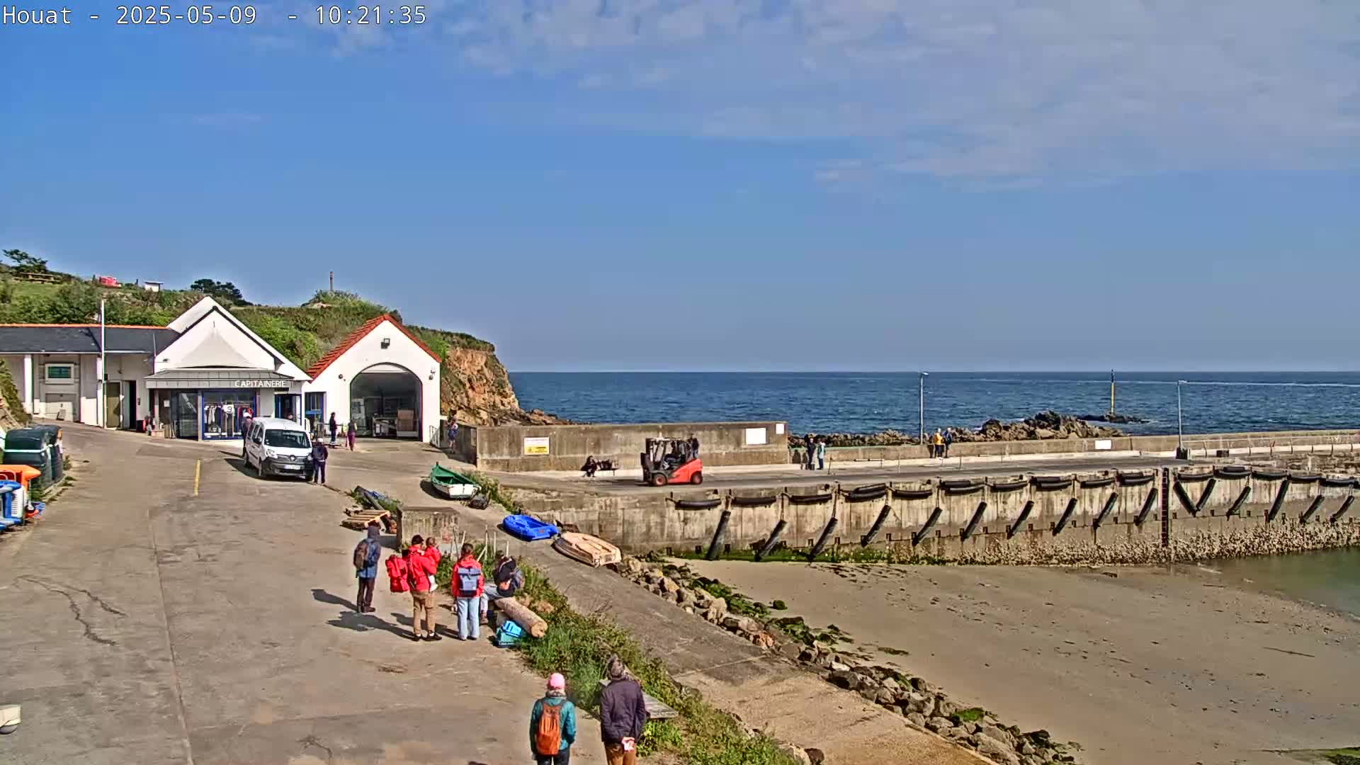 A sunny day at a harbor shows a small building, a few people, a forklift, some boats, and a calm ocean.