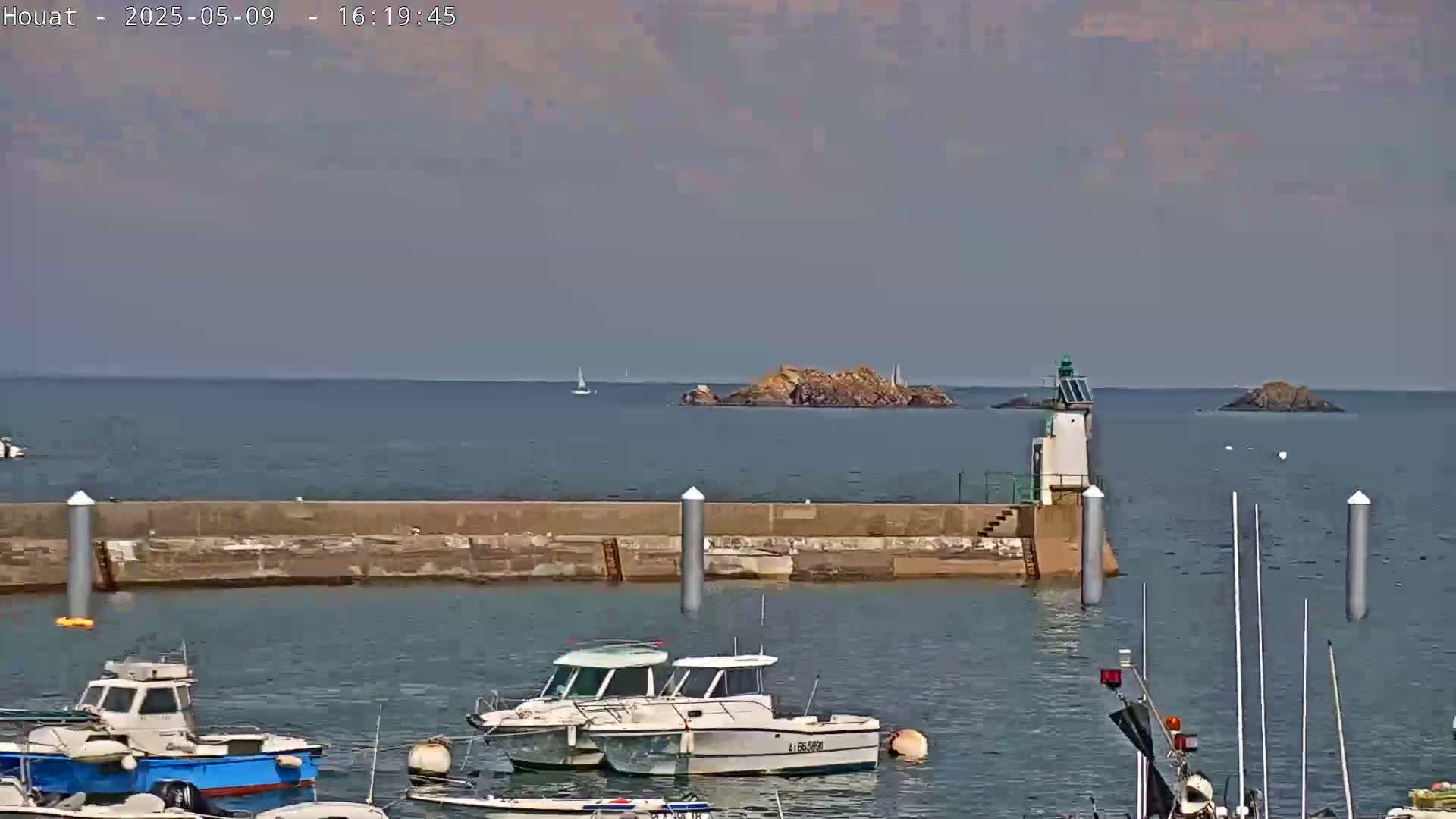 A harbor with several small motorboats is seen, a stone seawall in the foreground, and a small island with a lighthouse visible in the distance under a mostly cloudy sky.