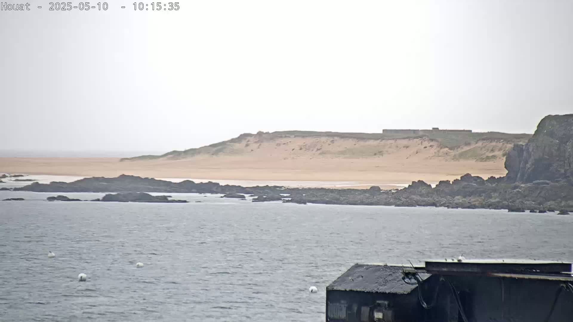 A calm, overcast day shows a sandy beach backed by a low, grassy hill, with dark rocks in the foreground separating the beach from a body of water.