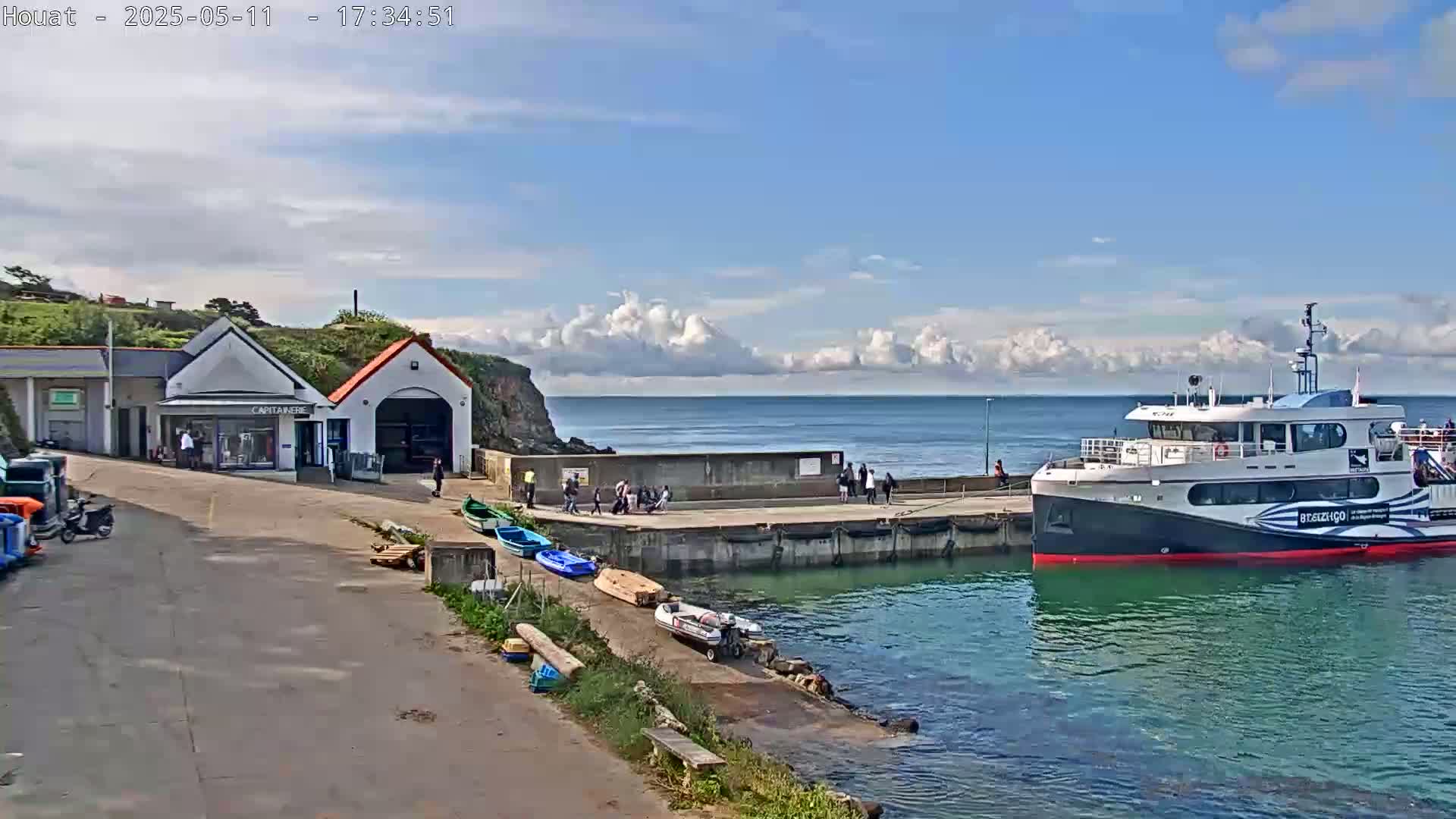 A ferry is docked at a harbor on a partly cloudy day, with several small boats and people near a small building.