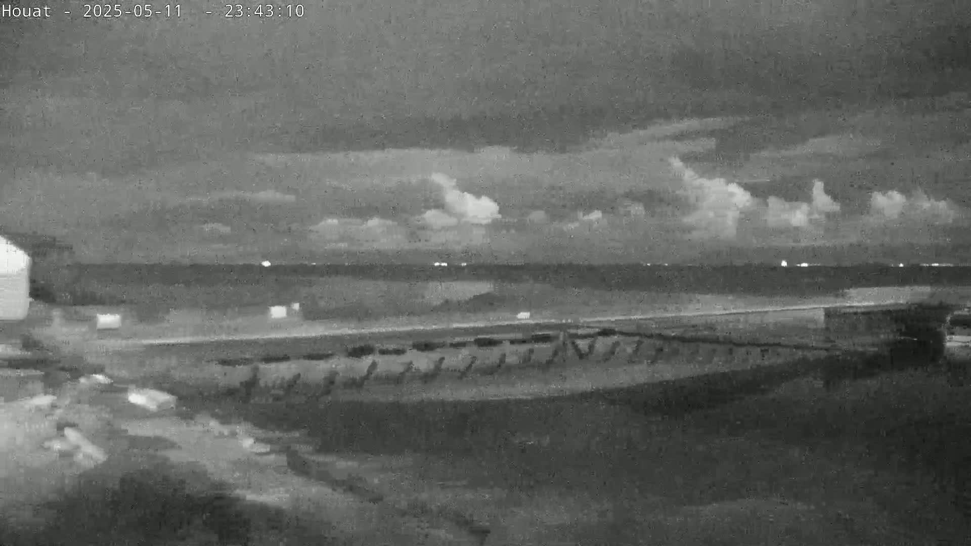 A nighttime, overcast view of a flat, dark landscape near a body of water with faint lights visible on the horizon.