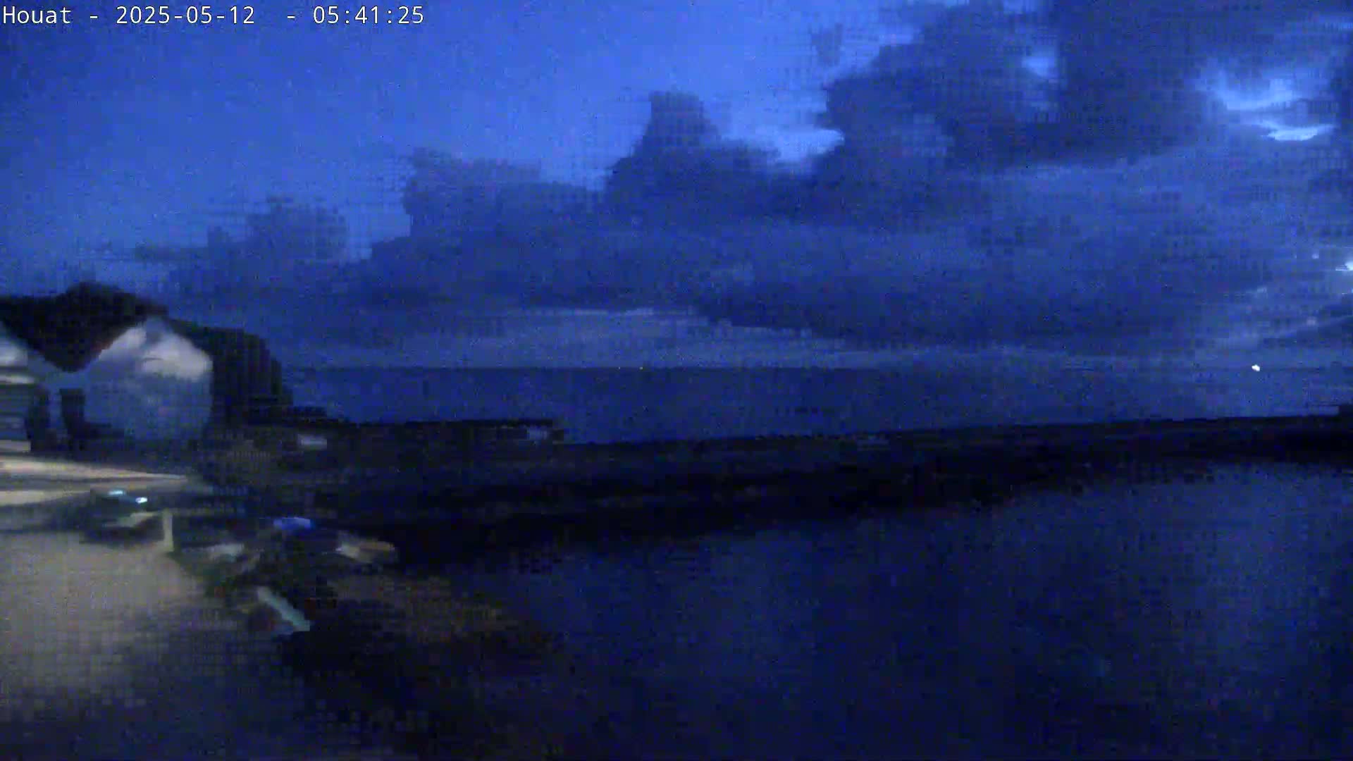 A dark-blue, twilight sky with clouds hangs over a calm body of water next to a dark shoreline and a building.