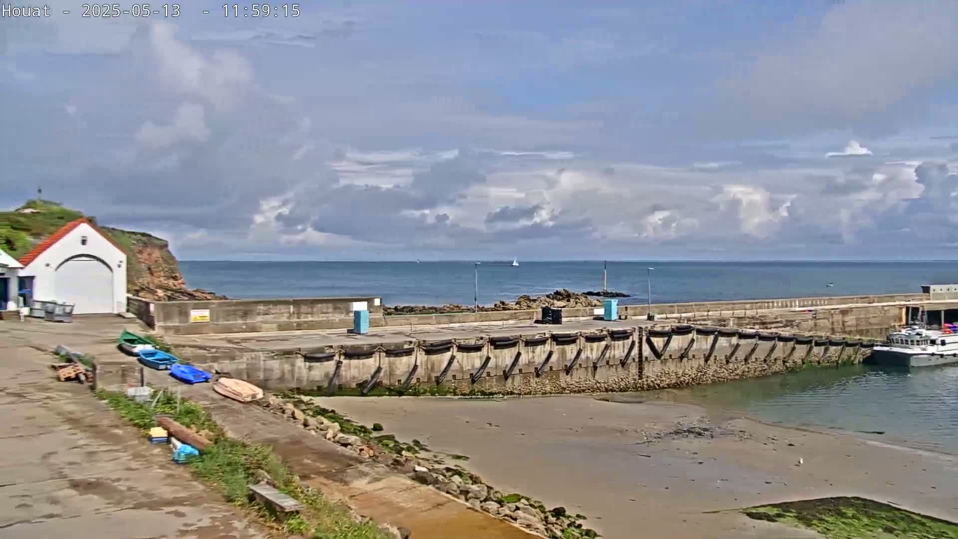 A small harbor with a few boats, a seawall, and a building is seen on a partly cloudy day.