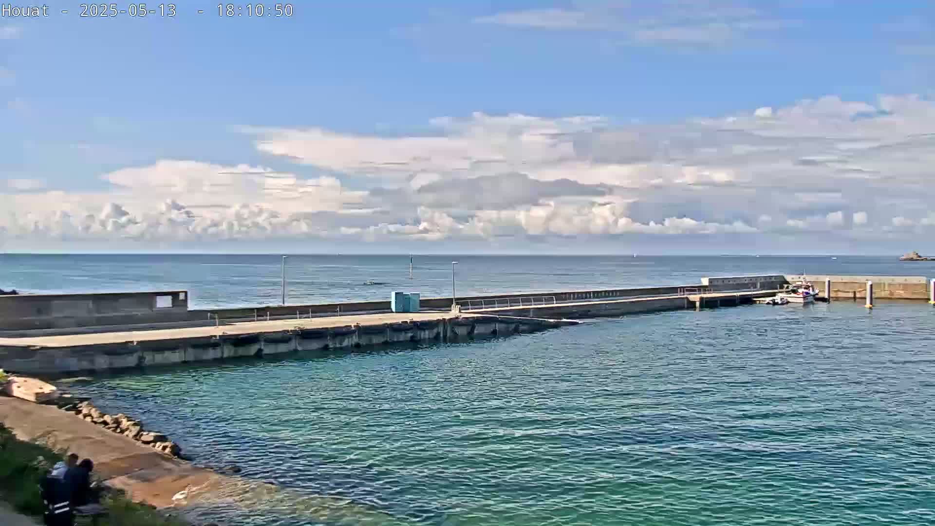 A partly cloudy day reveals a small harbor with a concrete pier and a few boats, the calm water reflecting the blue sky.